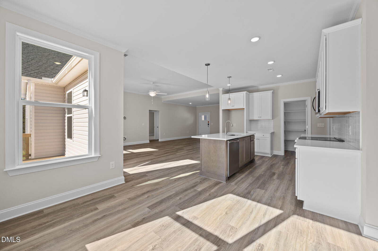 A bright, modern open-concept kitchen and living area with light wood floors, white cabinets, a large island, and pendant lights. Sunlight streams in through a window, creating a welcoming atmosphere.