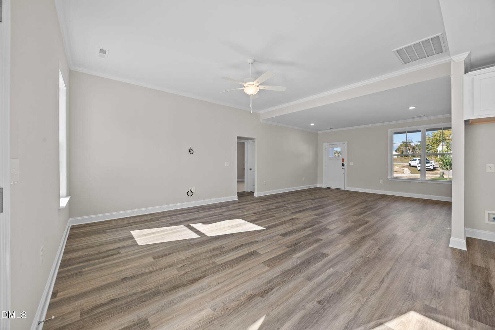 Bright, empty room with light gray walls, wood flooring, ceiling fan, sunlight through windows, and open layout leading to front door and kitchen area.