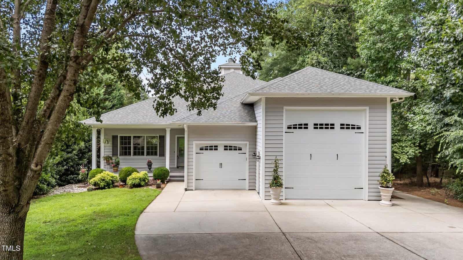 A single-story gray house with a front porch, two white garage doors, potted plants, manicured shrubs, and a large driveway, surrounded by green trees and lawn.