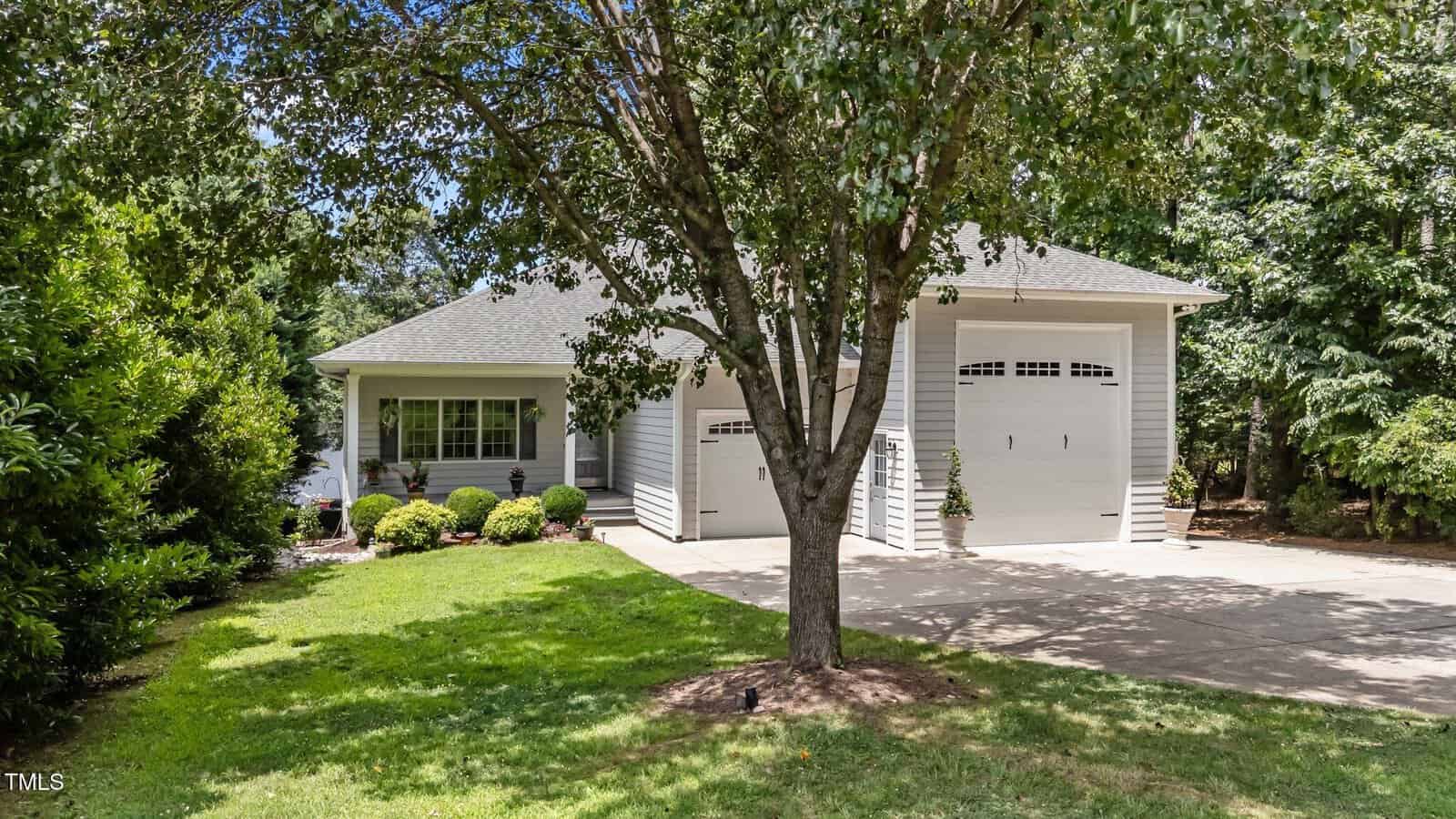 A single-story house with a gray roof, white siding, large double garage doors, a well-kept lawn, shrubs, and a tree in the front yard surrounded by greenery.