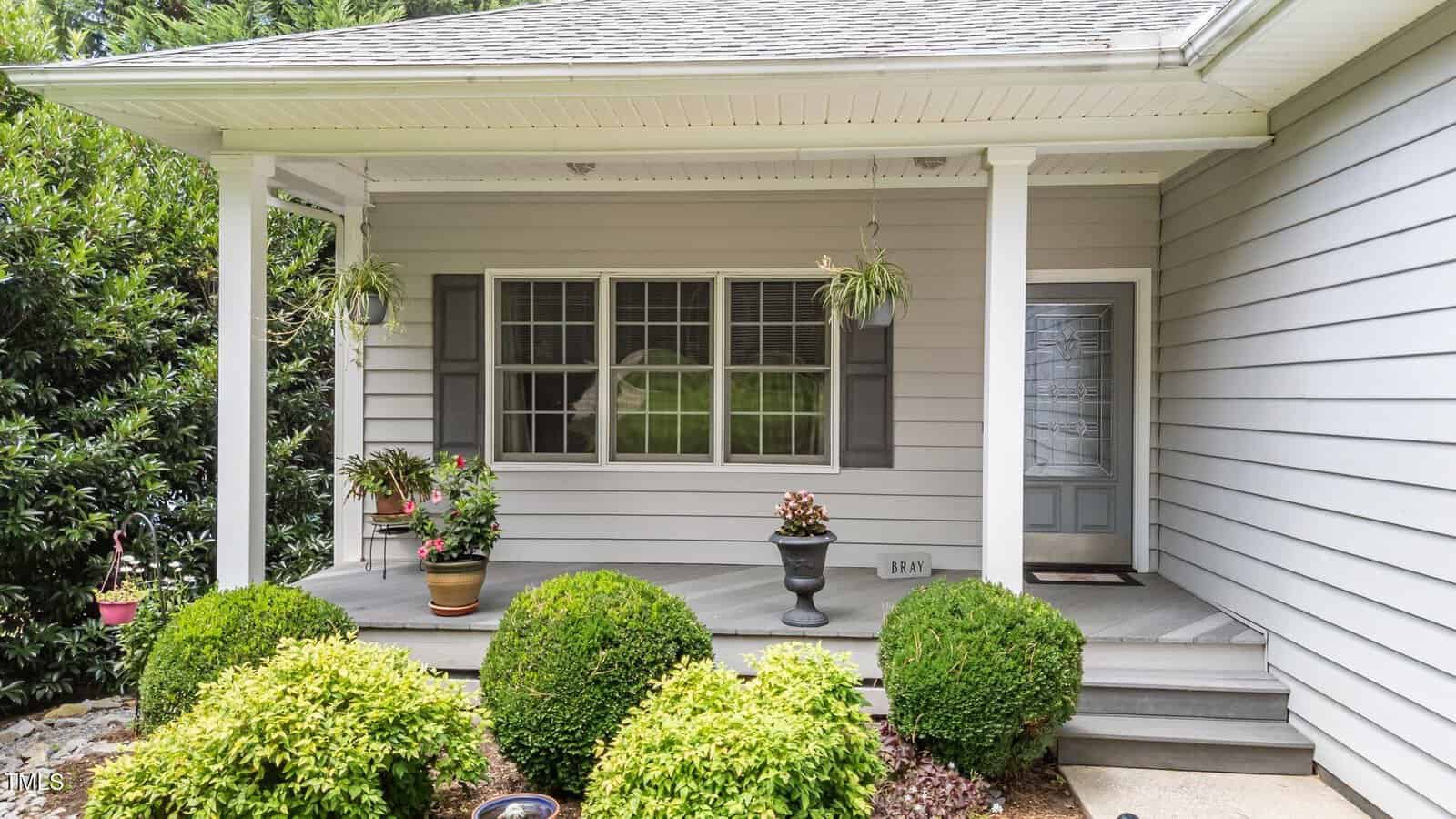A gray house with white trim features a covered front porch, potted plants, hanging baskets, and a gray front door. Bushes and landscaping fill the foreground.
