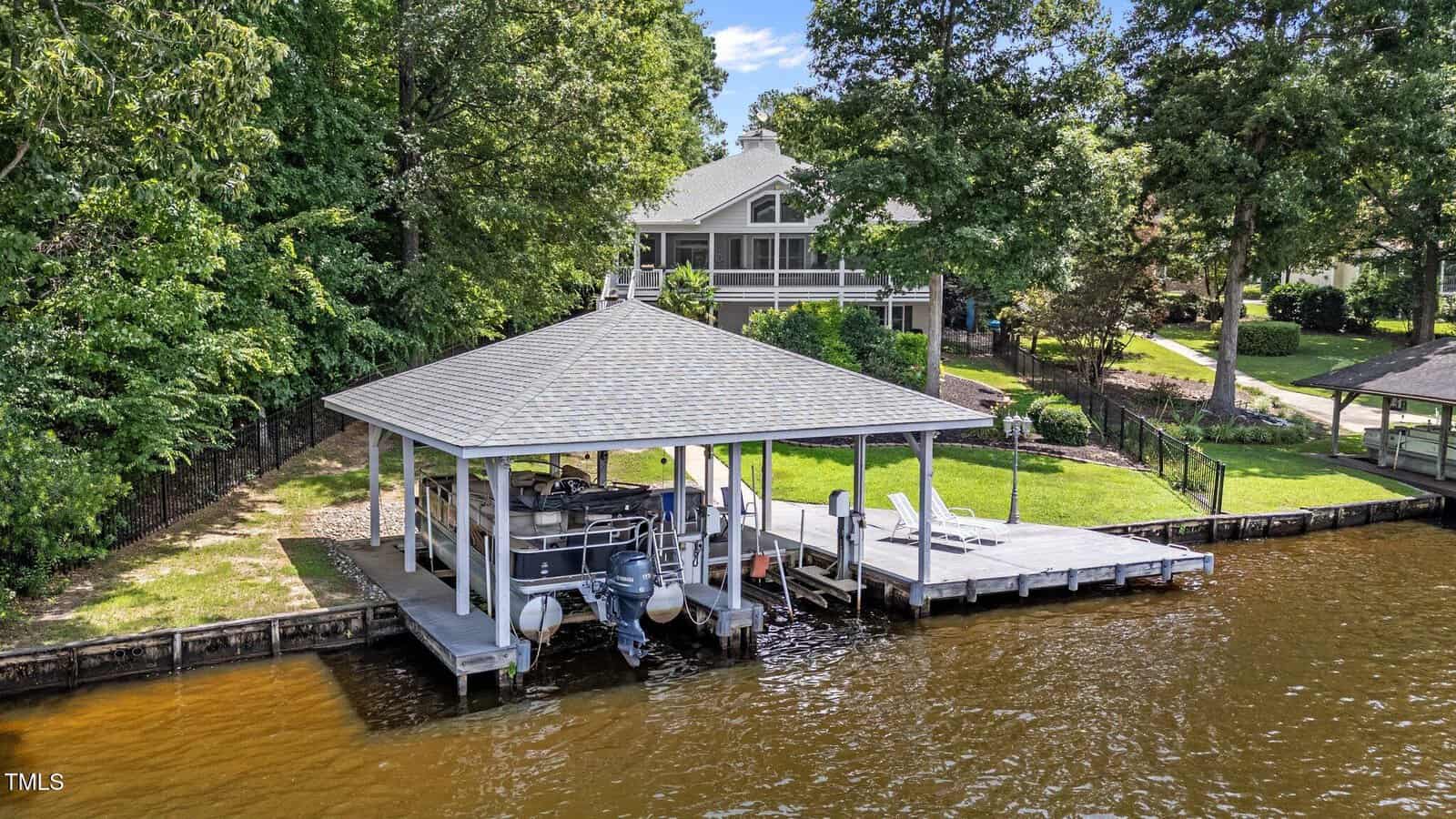A covered boat dock with a boat and seating area sits on a calm brown lake. The dock is next to a house surrounded by trees and greenery. The shoreline has a wooden bulkhead and a grassy yard leading up to the house.