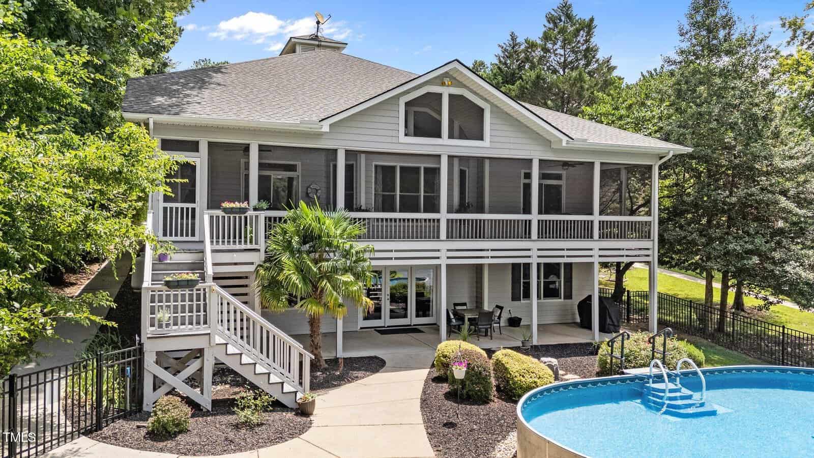 Two-story gray house with a large screened porch, white railings, and outdoor stairs, facing a backyard with a round above-ground pool, patio seating area, and landscaped bushes surrounded by trees.