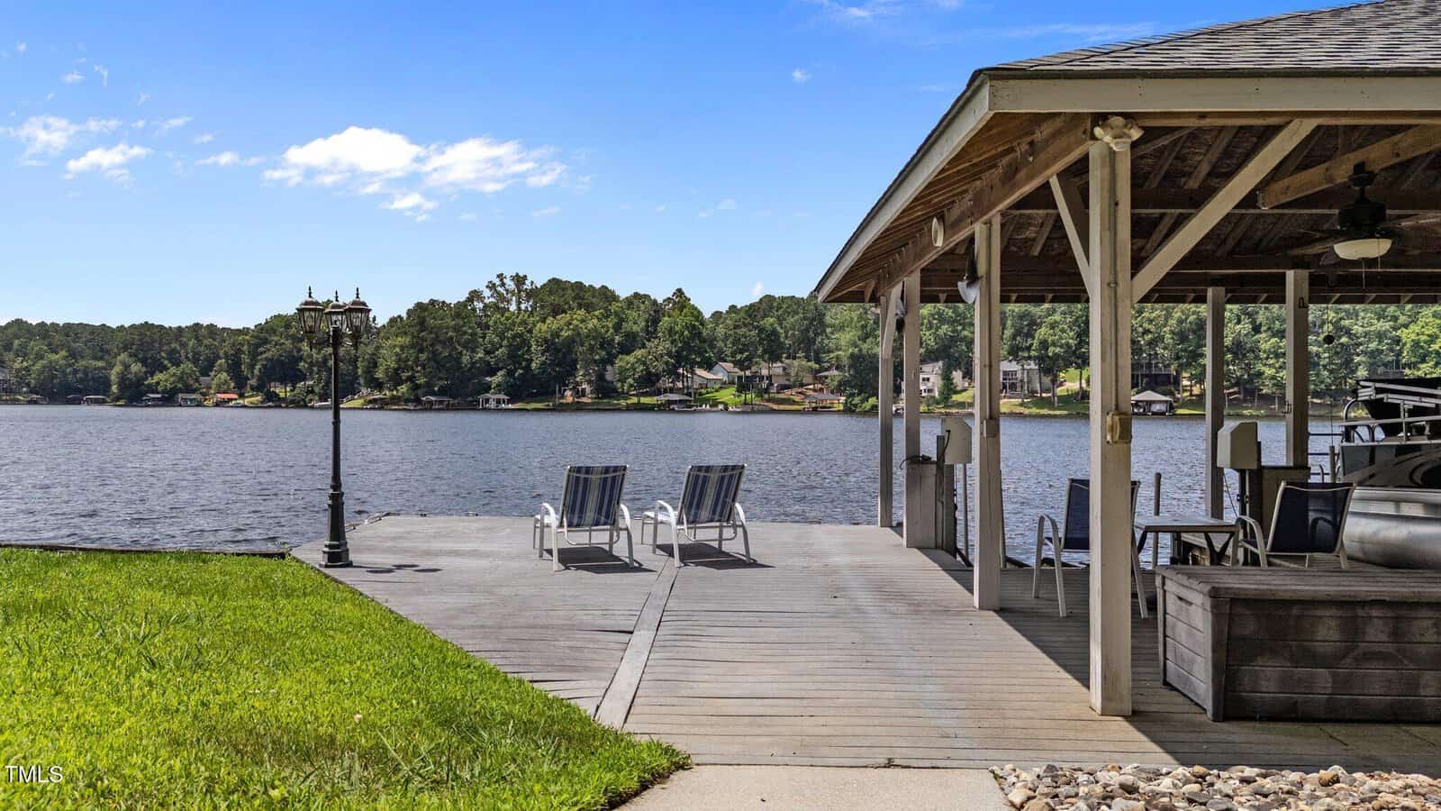 A wooden dock with two lounge chairs faces a calm lake. A covered area with seating and a boat is on the right. Green grass and trees surround the water under a bright blue sky with scattered clouds.