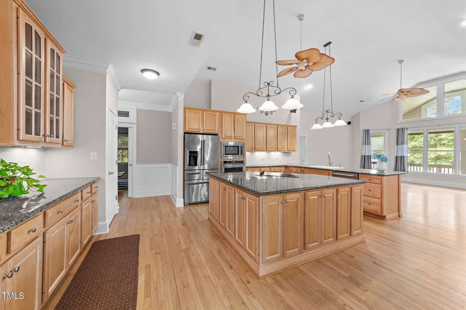 Spacious kitchen with light wood cabinets, island with granite countertop, stainless steel appliances, and hardwood floors. Ceiling fans and pendant lights hang above. Large windows in the adjacent living area let in natural light.
