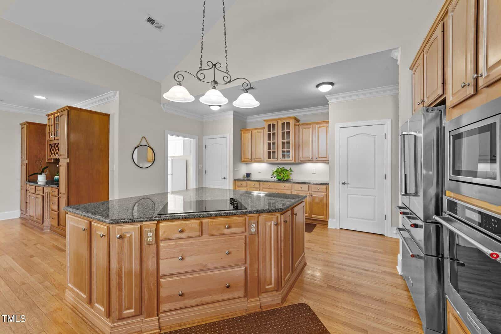 Spacious kitchen with wood cabinets, a large central island with a granite countertop, stainless steel appliances, hardwood floors, and pendant lighting overhead. A closed white door is visible at the back.