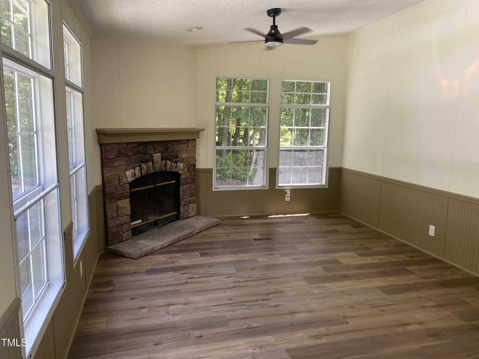 A room with wood flooring, beige wainscoting, and white walls. It has large windows, a ceiling fan, and a corner stone fireplace with a hearth. Sunlight comes through the windows, revealing a wooded view outside.