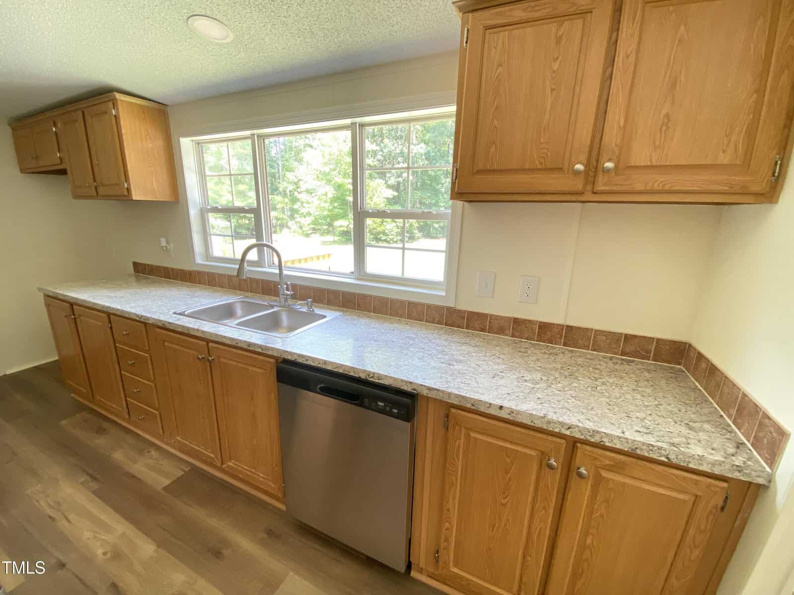 A kitchen with wooden cabinets, a speckled granite countertop, a stainless steel double sink, a dishwasher, tile backsplash, and large windows letting in natural light.