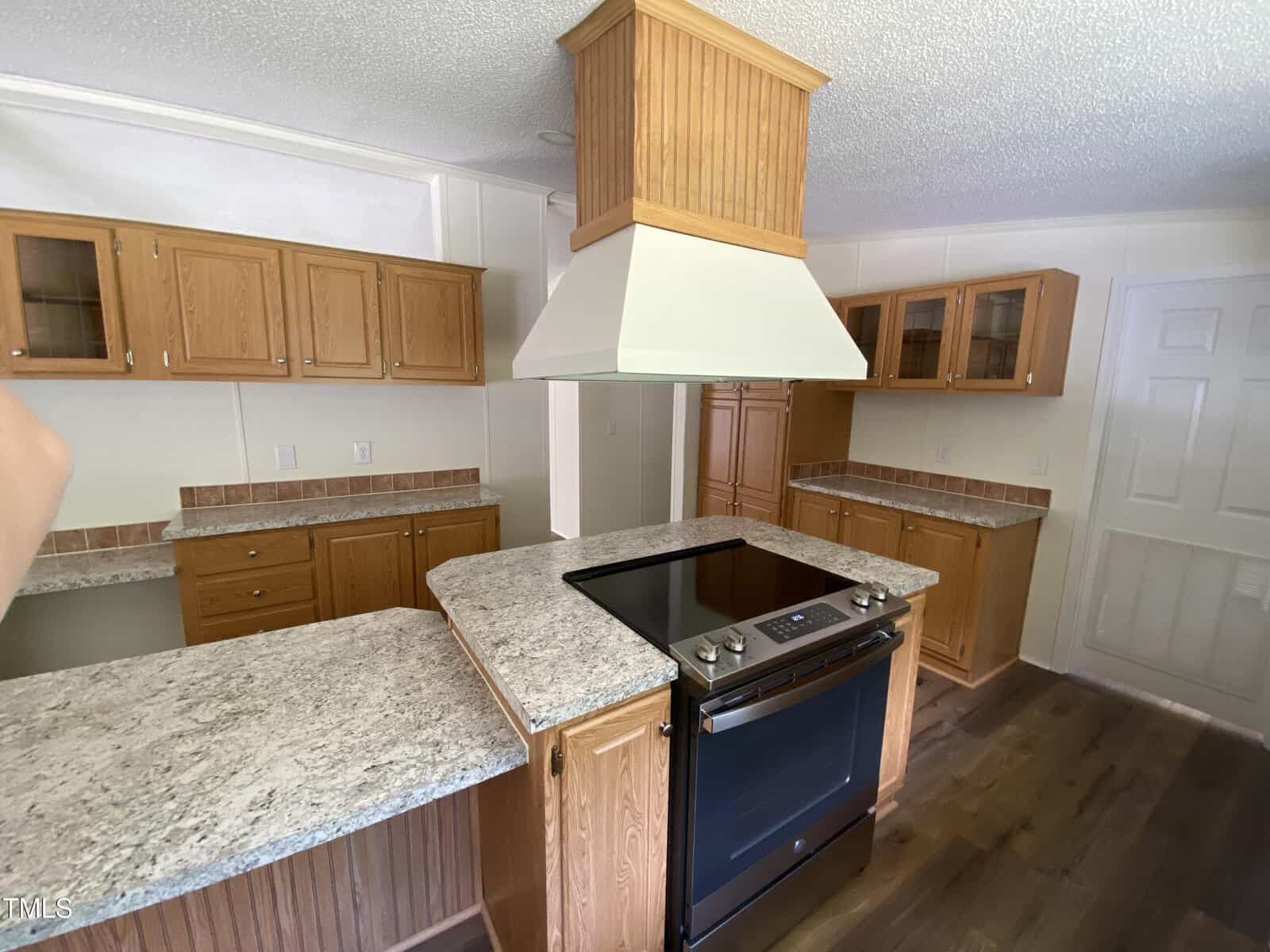 A kitchen with wood cabinets, granite-style countertops, an island with a stovetop and oven, a large range hood, and wood flooring. Upper and lower cabinets provide storage, and the walls are light-colored.