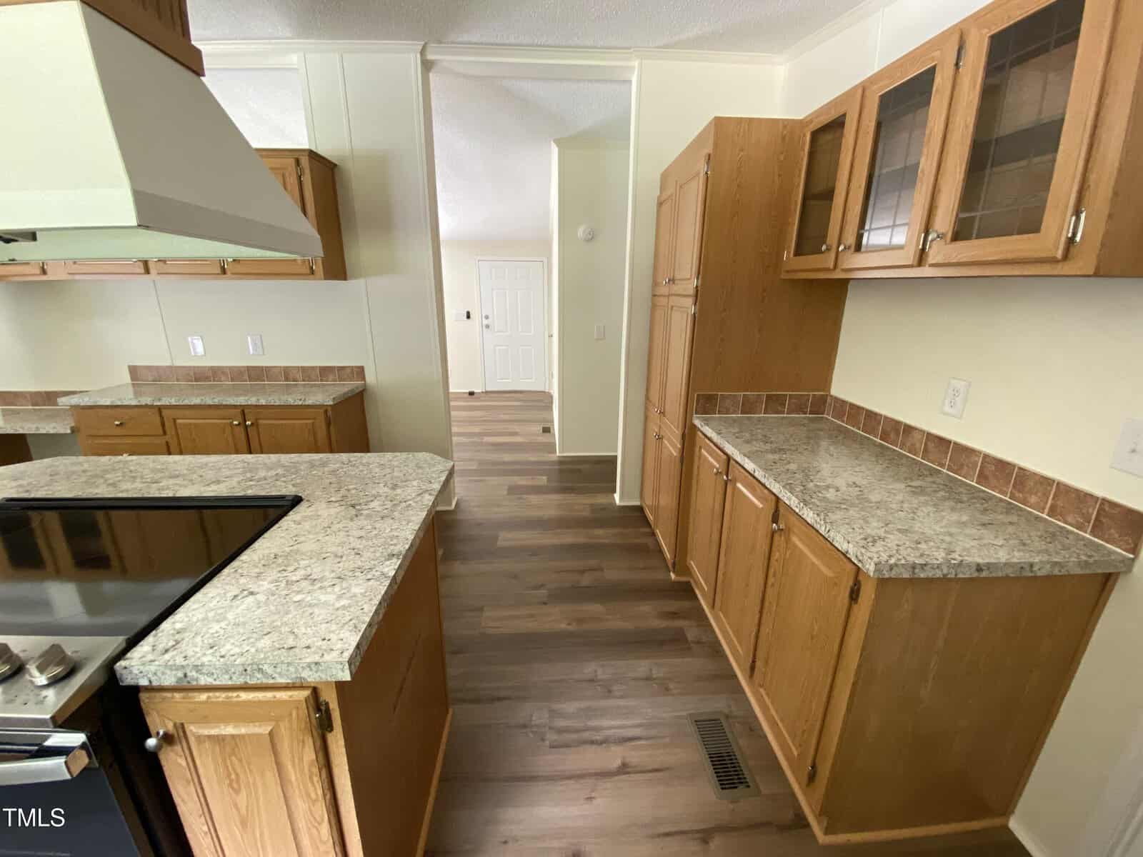 A kitchen with wood cabinets, granite-style countertops, a stove, and vent hood. The floor is wood laminate, and there is a tiled backsplash. An open doorway leads to a white front door in the background.