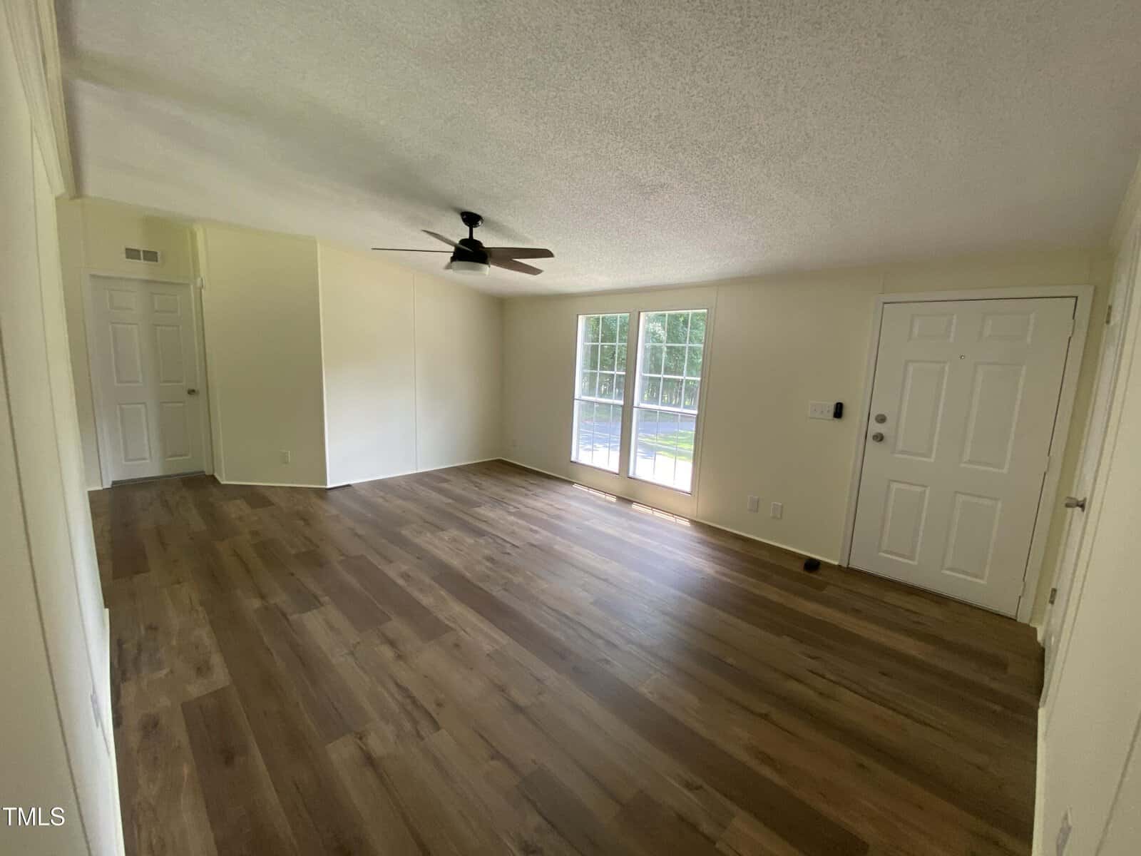 An empty room with light-colored walls, wood-style flooring, a ceiling fan, two large windows, and a white front door. Natural light enters through the windows, illuminating the spacious area.