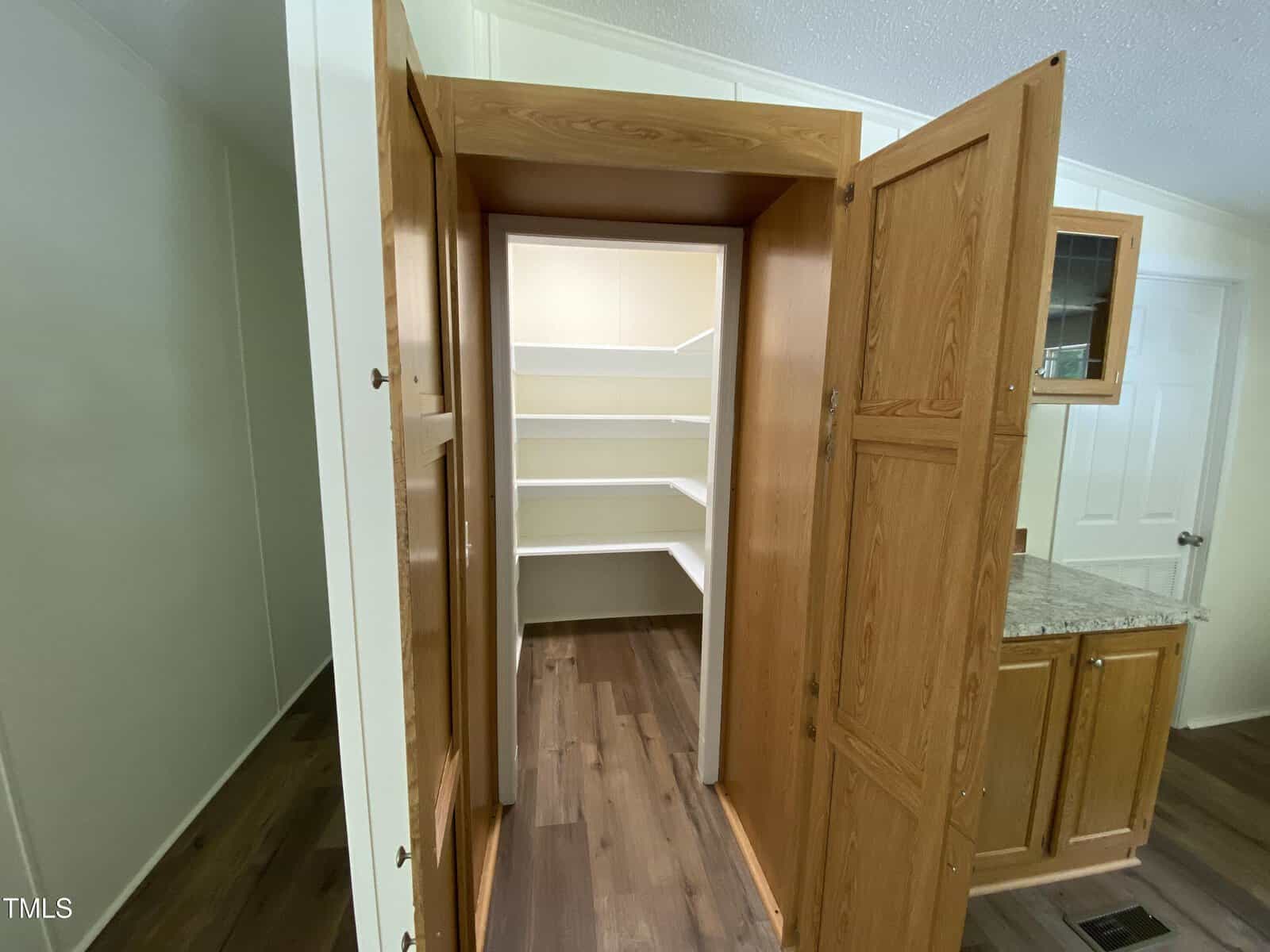 A wooden pantry with open double doors reveals a walk-in space with white built-in shelves. To the right, a kitchen counter with a granite surface and cabinet is partially visible.