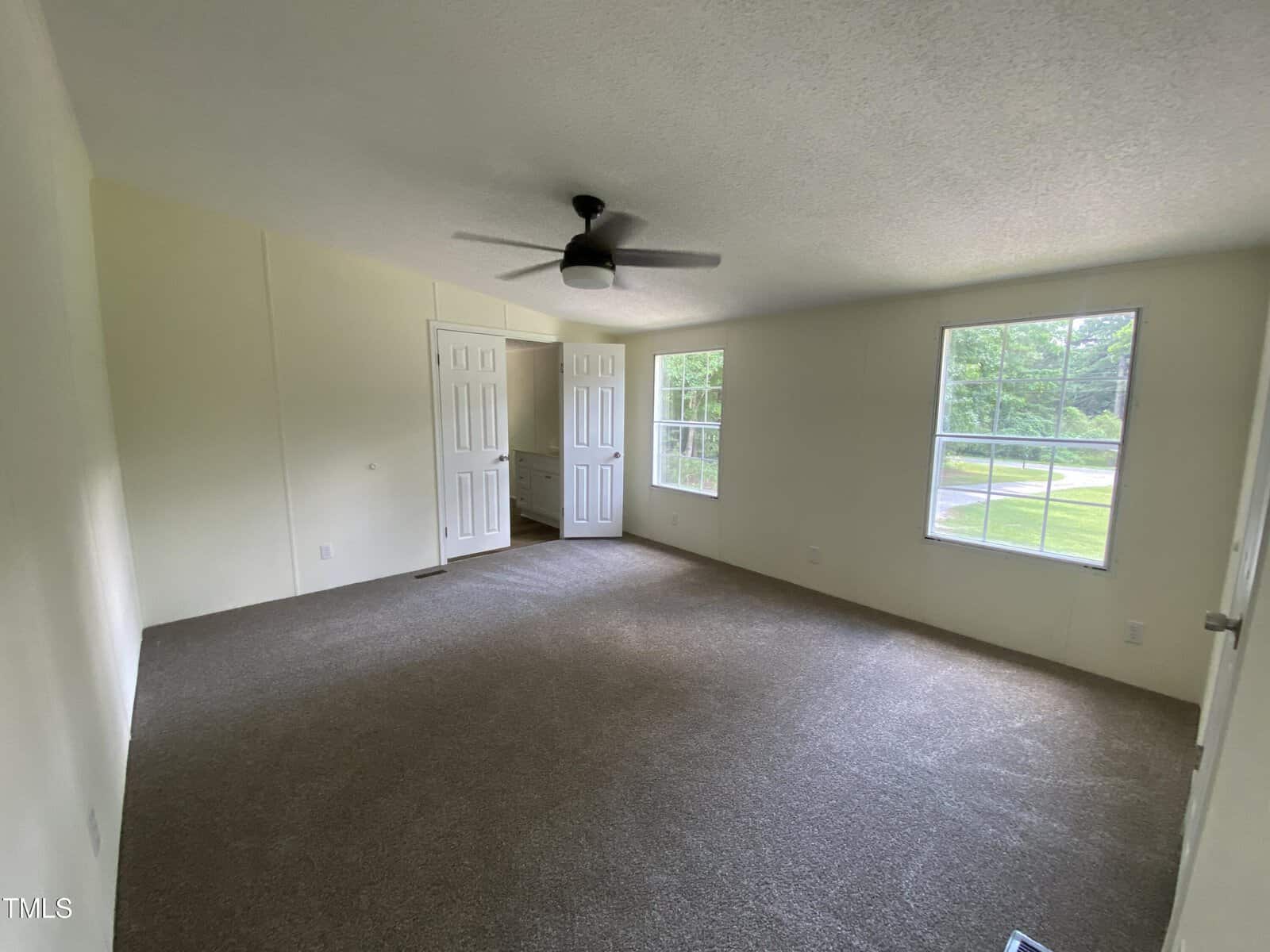 A carpeted, empty room with light-colored walls, a ceiling fan, two windows letting in natural light, and double doors leading to another area. The room appears clean and spacious.