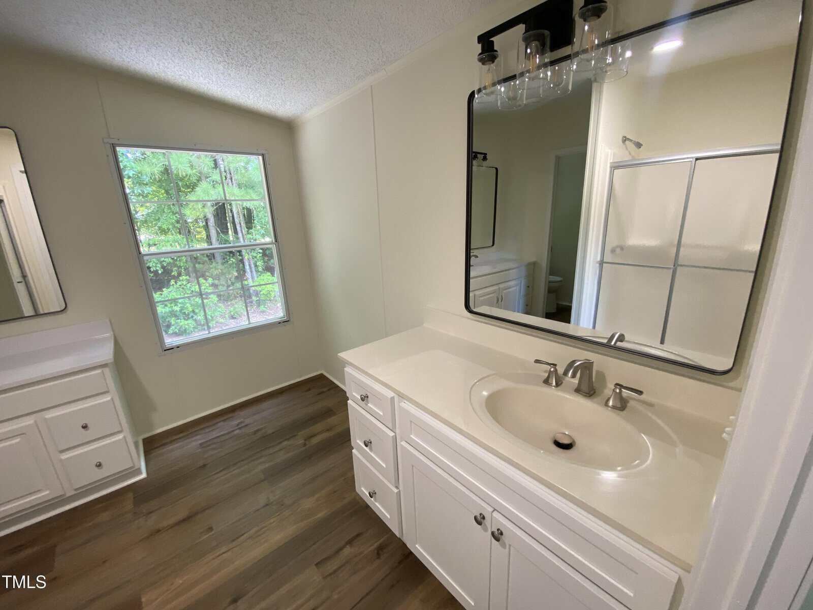 Bright bathroom with white vanity, sink, and mirror, wood-style flooring, large window with outdoor view, and a shower with glass doors in the background.