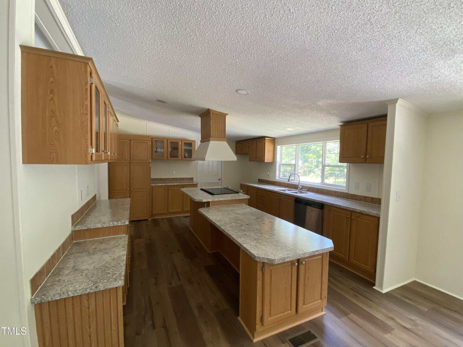 A spacious kitchen with wood cabinets, stone countertops, a center island, stovetop with range hood, double sink under large windows, and wood flooring. The space is well-lit with natural light from the windows.