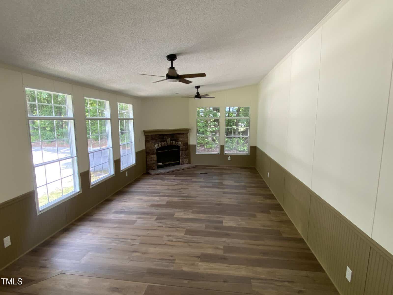 Bright, empty room with wood floors, ceiling fans, large windows on one side, and a stone fireplace in the corner. The walls are painted cream and olive green, with natural light streaming in.