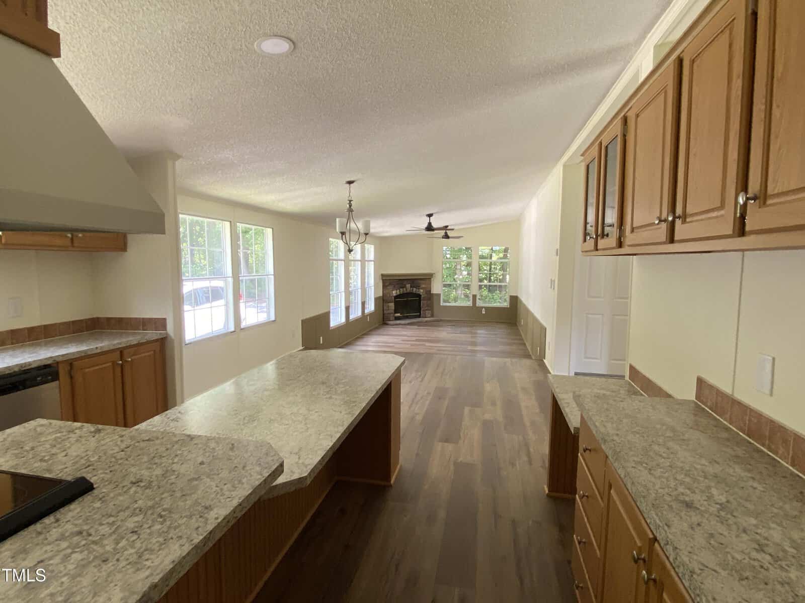 A view of an open-concept kitchen and living area with wood cabinets, marble-style countertops, and wood floors. The space has large windows, a ceiling fan, and a fireplace at the far end of the living room.