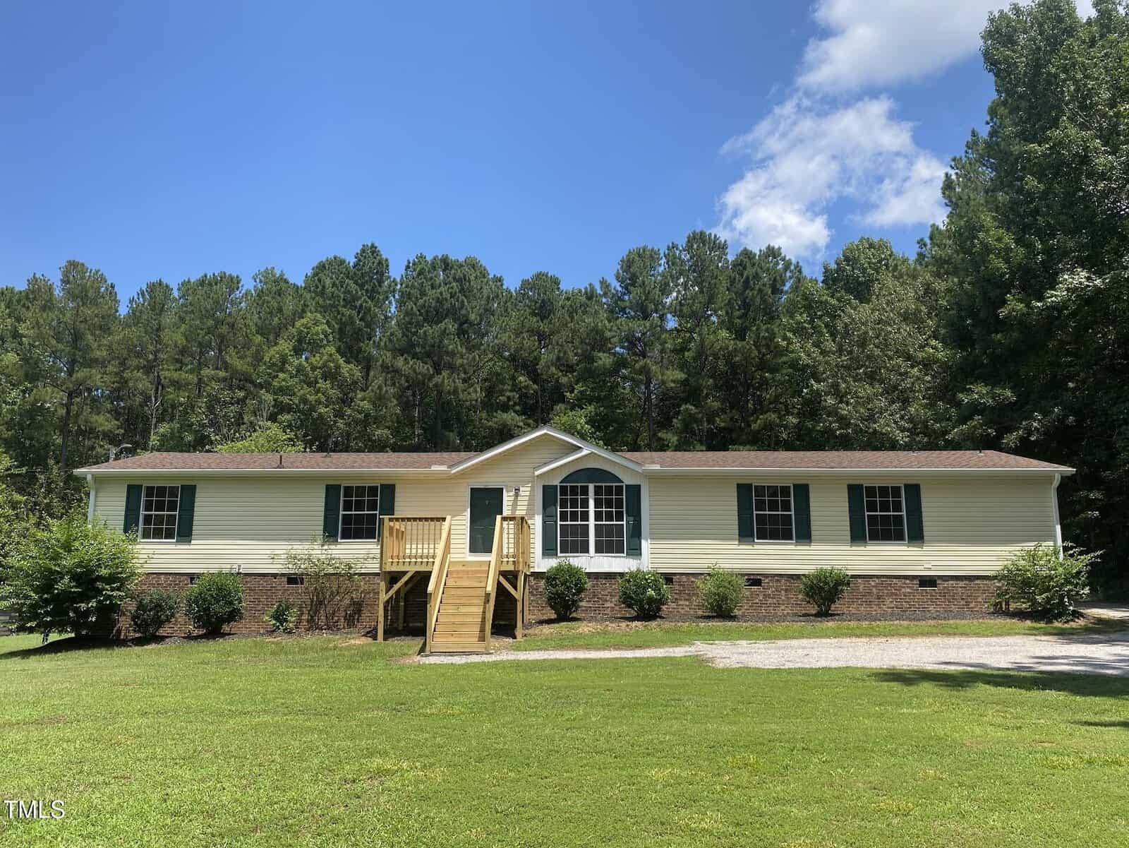 Single-story manufactured home at 4037 Crestview Lane in Oxford features a brick foundation, beige siding, and brown roof. Wooden steps lead to the front door, with a grassy lawn, shrubs, and trees under a partly cloudy blue sky.