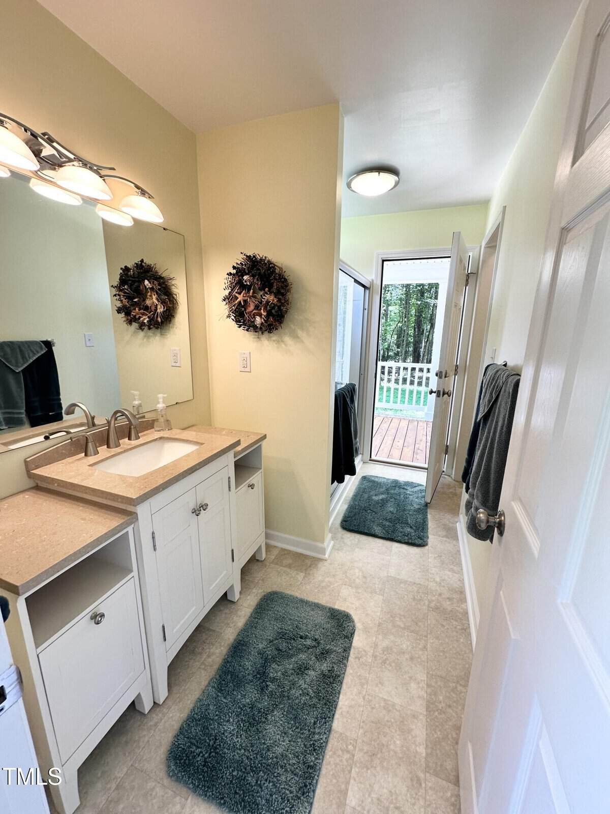 A bright bathroom with a double sink vanity, two wreaths above each sink, green bath mats, towel racks, and an open door leading to a deck with a wooded view outside.