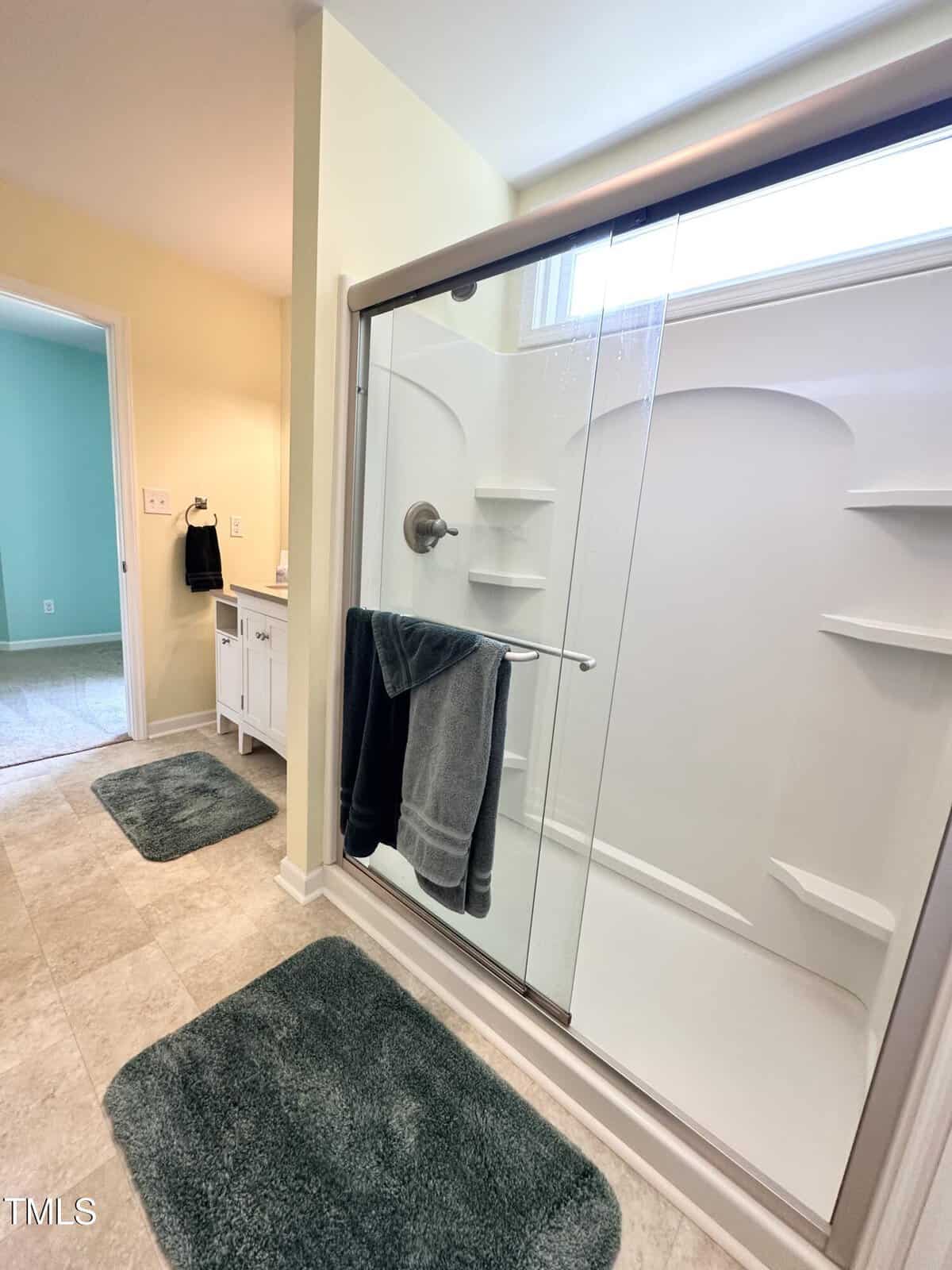 Modern bathroom with a glass-enclosed shower, two dark towels on the shower door, white vanity, and green bath mats on tiled floor. Light yellow walls and an adjacent room visible through the doorway.