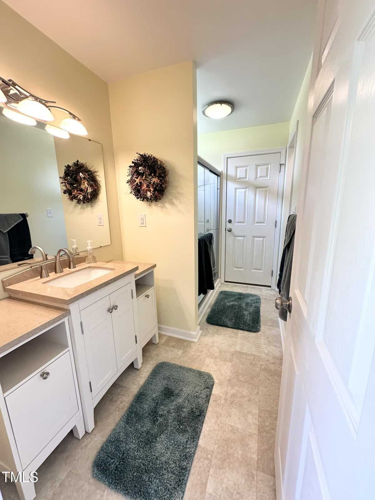 A bright bathroom with double sinks, white cabinetry, two green rugs, a glass shower, a wreath on the mirror, and a door at the end of the room. Warm lighting creates a clean and welcoming atmosphere.