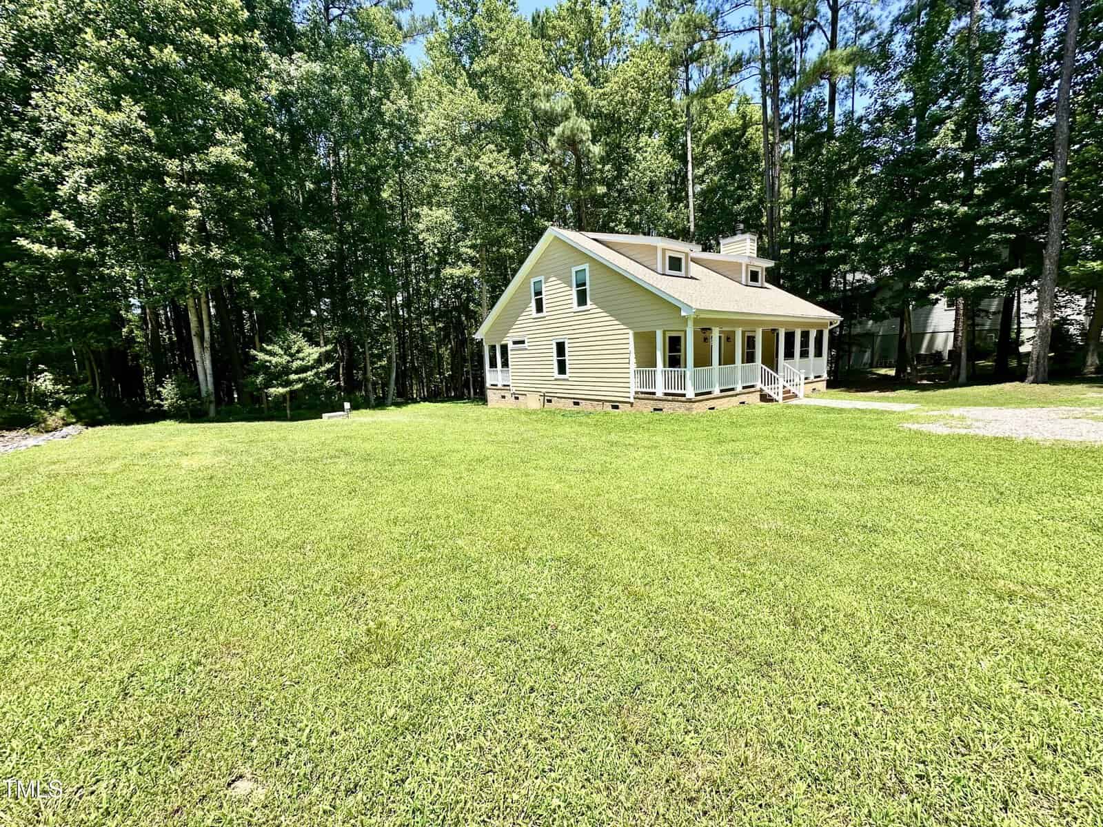A tan two-story house with a covered front porch sits on a large, green lawn bordered by tall trees. The house has white trim and multiple windows, and the sky is clear and sunny.
