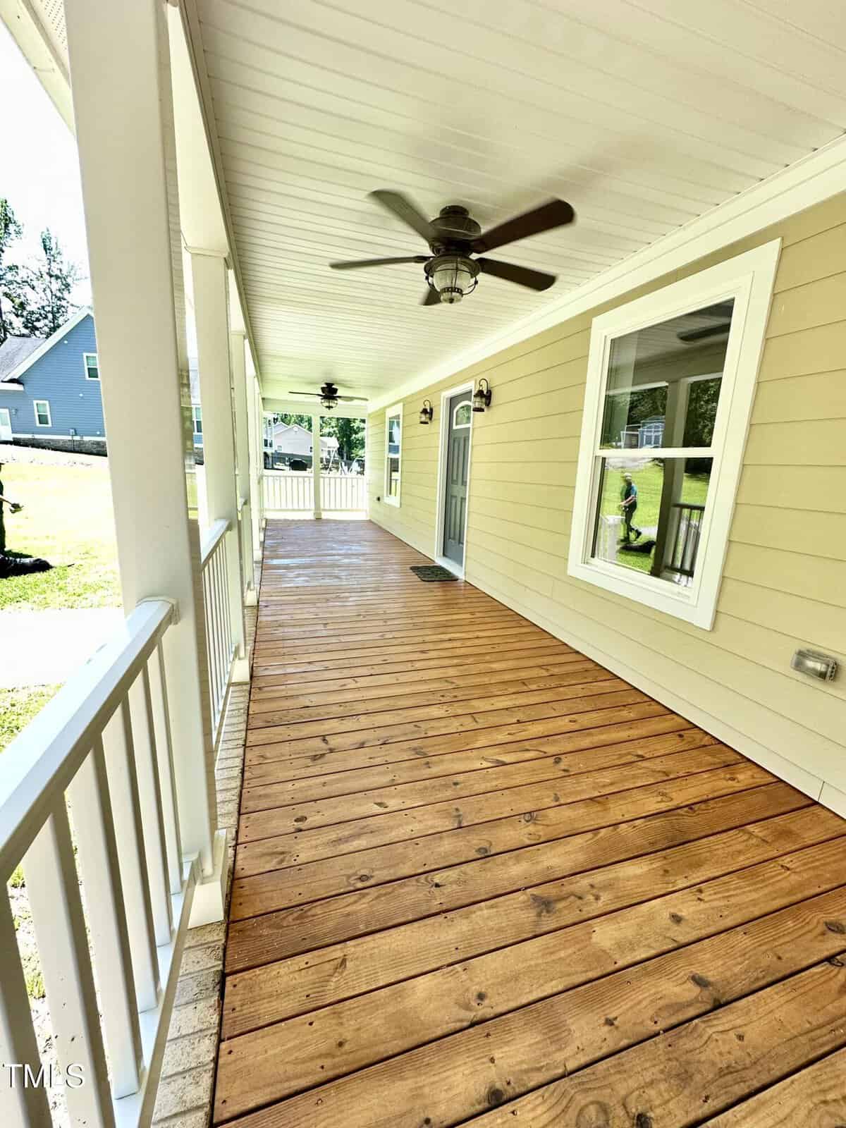A covered wooden porch with a ceiling fan, white railing, and beige siding. A gray door and white-framed window are visible, with a grassy yard and another house in the background.