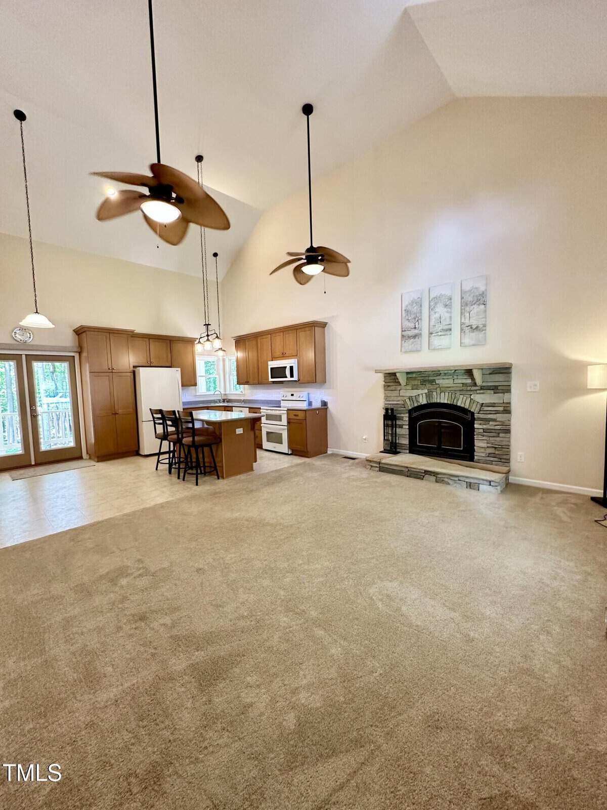 Spacious open-concept living area with beige carpet, a stone fireplace, ceiling fans, and a kitchen featuring wood cabinets, an island, and white appliances. Three framed artworks hang above the fireplace.