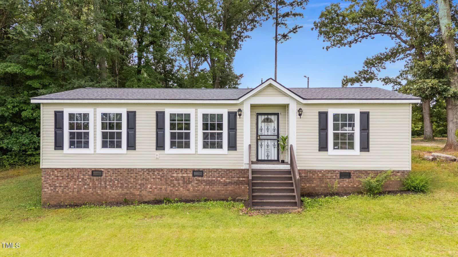 A small, single-story house with beige siding, black shutters, a brick foundation, and a front door with steps leading up. The house is surrounded by green grass and trees in the background.
