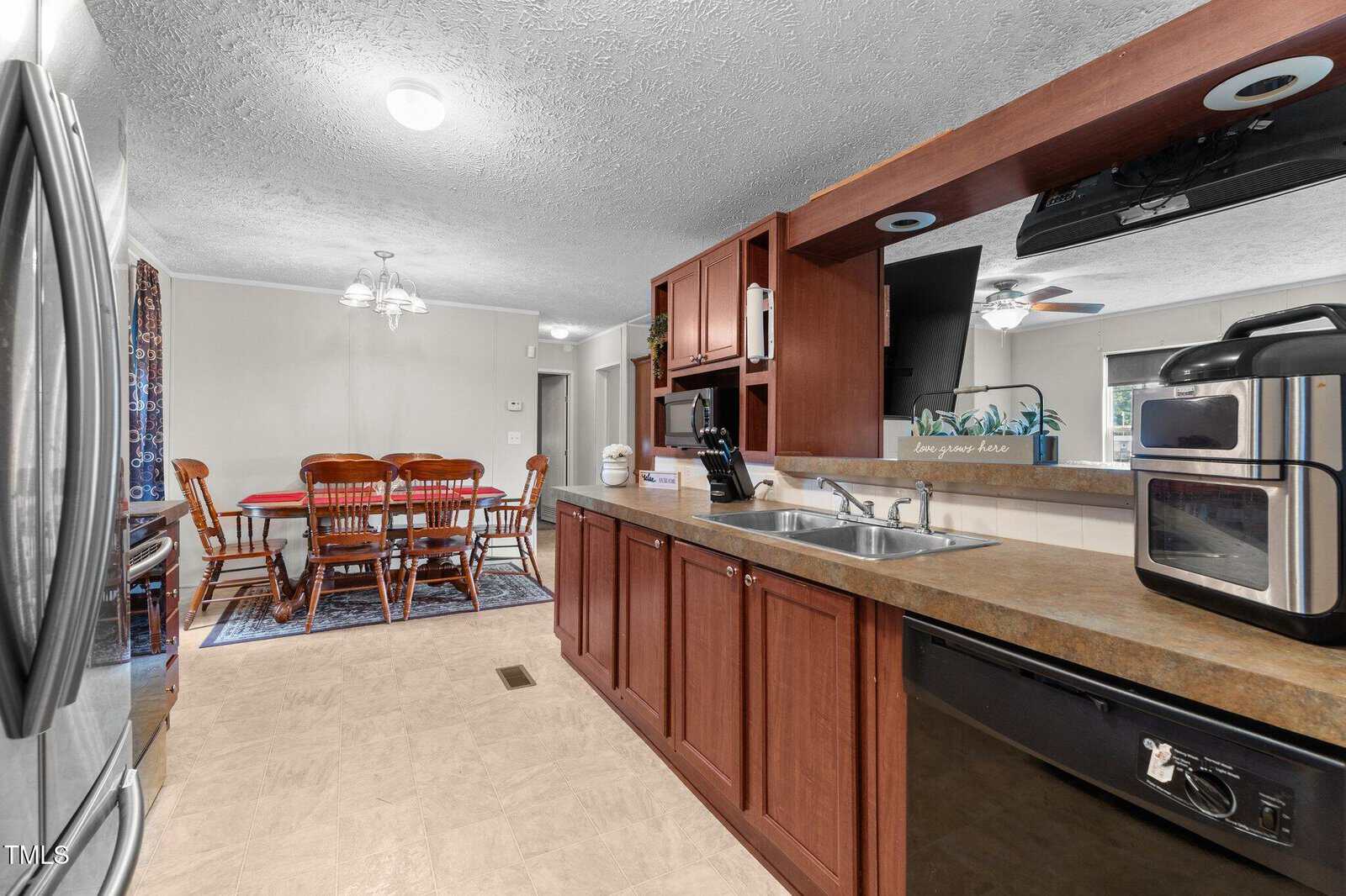 A kitchen with brown cabinets and a double sink, featuring a countertop with appliances. In the background, a dining area with a wooden table and six chairs is visible under a chandelier.