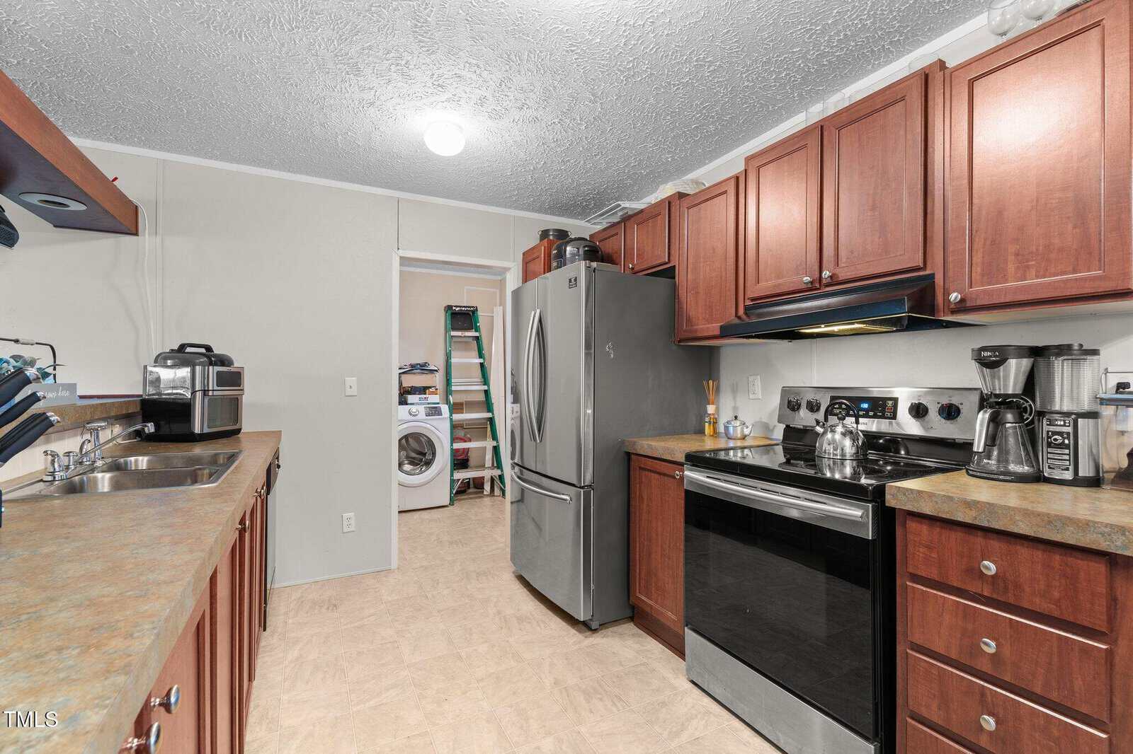 Kitchen with brown cabinets, stainless steel appliances, coffee maker, air fryer, and a view into a laundry room with a washer, dryer, ladder, and cleaning supplies. Countertops are clear with minimal items.