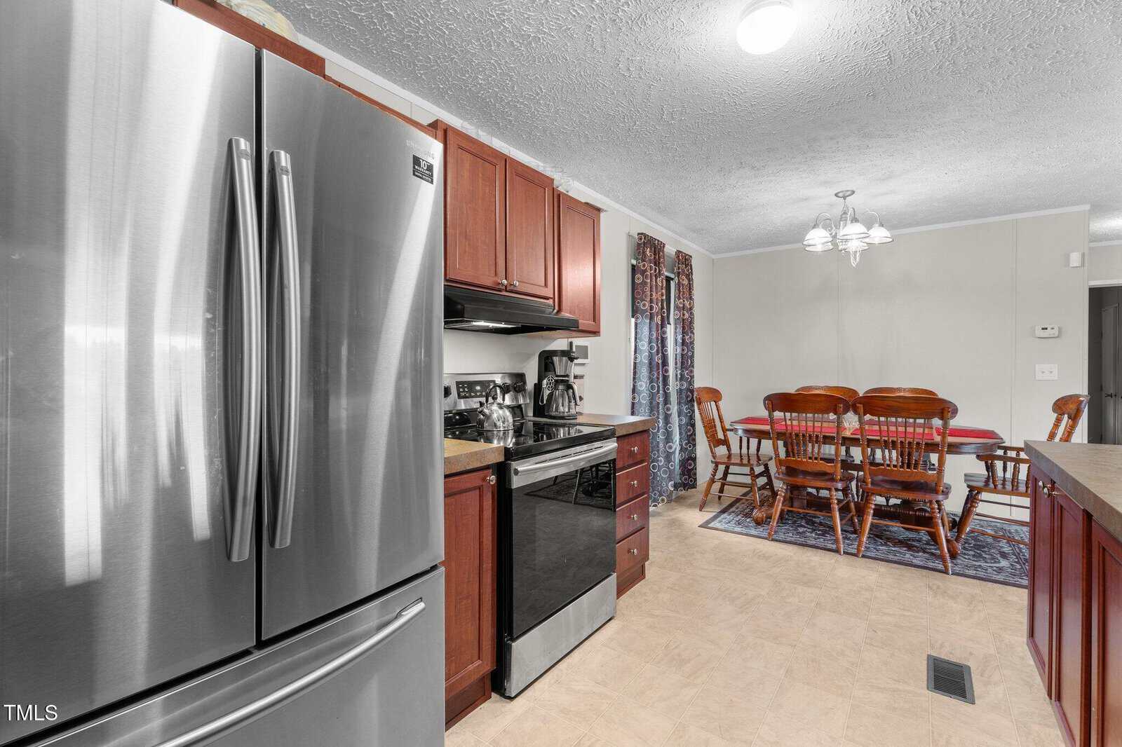 A kitchen with stainless steel fridge, stove, and microwave, dark wood cabinets, and a dining area with a wooden table and six chairs under a ceiling light. Patterned curtains cover the windows.
