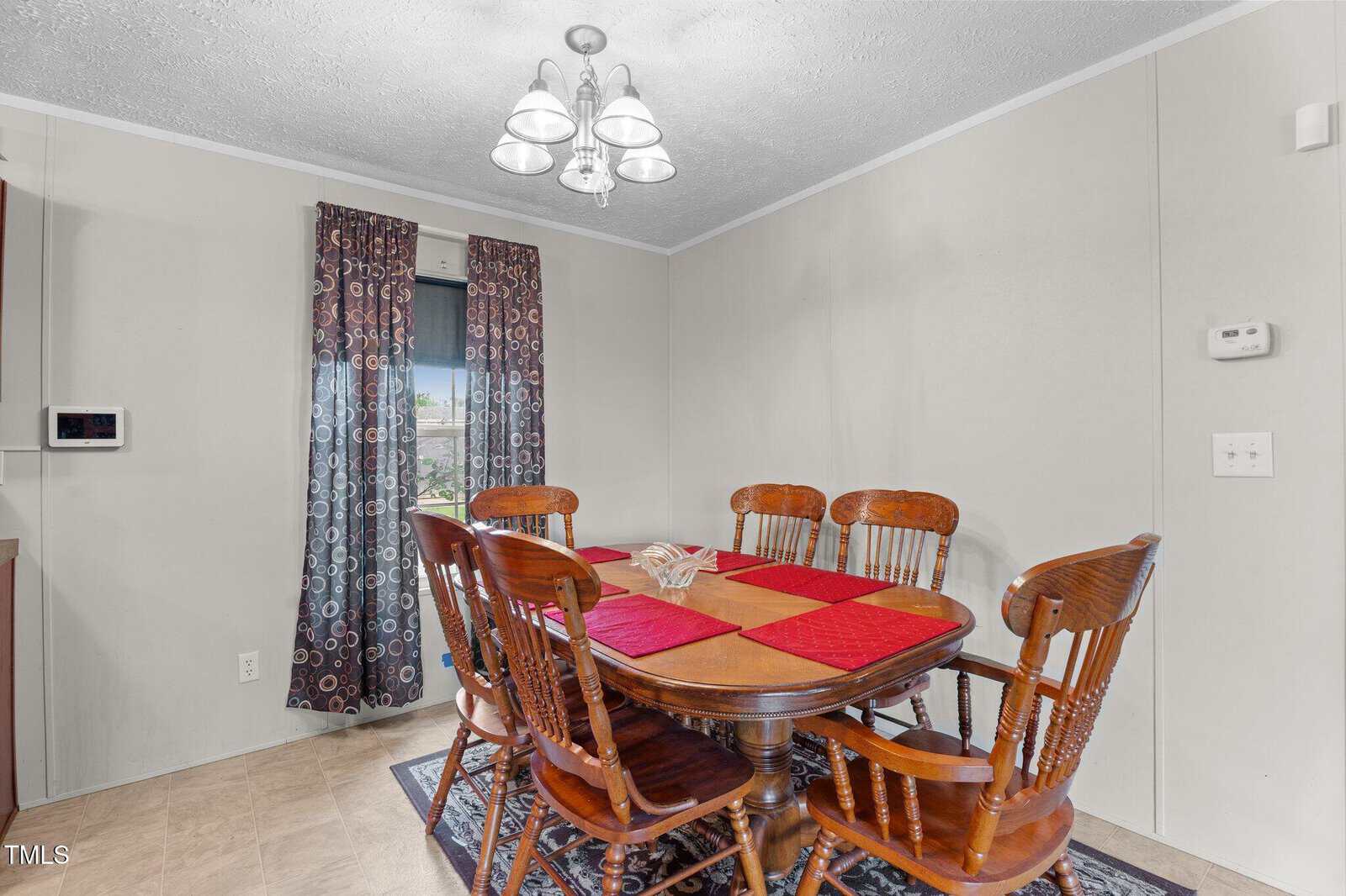 A dining area with a wooden table set for six, red and gold placemats, wooden chairs, a light fixture above, long patterned curtains, a window, and beige walls with a thermostat and light switch visible.