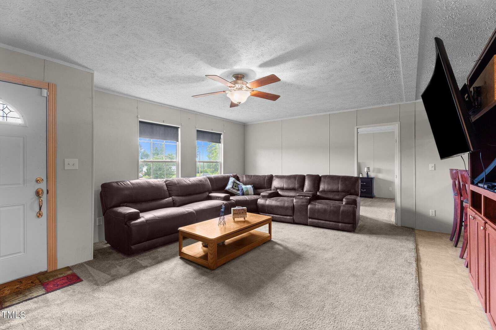 A spacious living room with a large L-shaped brown sectional sofa, a wooden coffee table, a ceiling fan, three windows with blue curtains, and a wall-mounted TV. The carpet is light beige, and there is a red cabinet on the right.