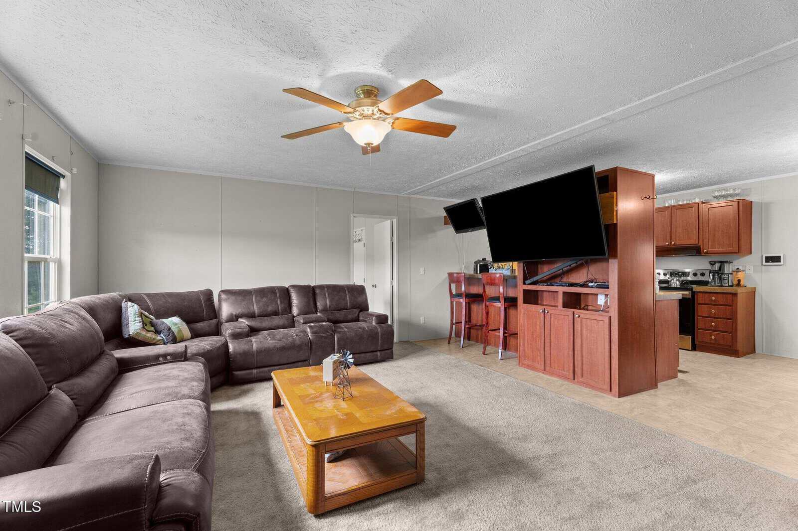 A spacious living room with brown sectional sofas, a wooden coffee table, beige carpet, a ceiling fan, a large wall-mounted TV, and an open view into a kitchen with wooden cabinets. Natural light comes in through a window.