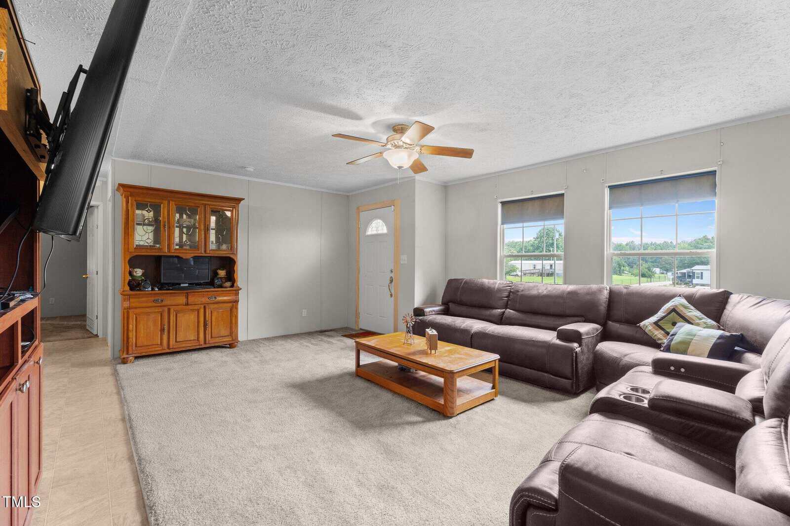 A living room with beige carpet, a brown sectional sofa, a wooden coffee table, and a wooden entertainment cabinet. Three windows with blue blinds and a ceiling fan are also visible. A flat-screen TV is mounted on the wall.