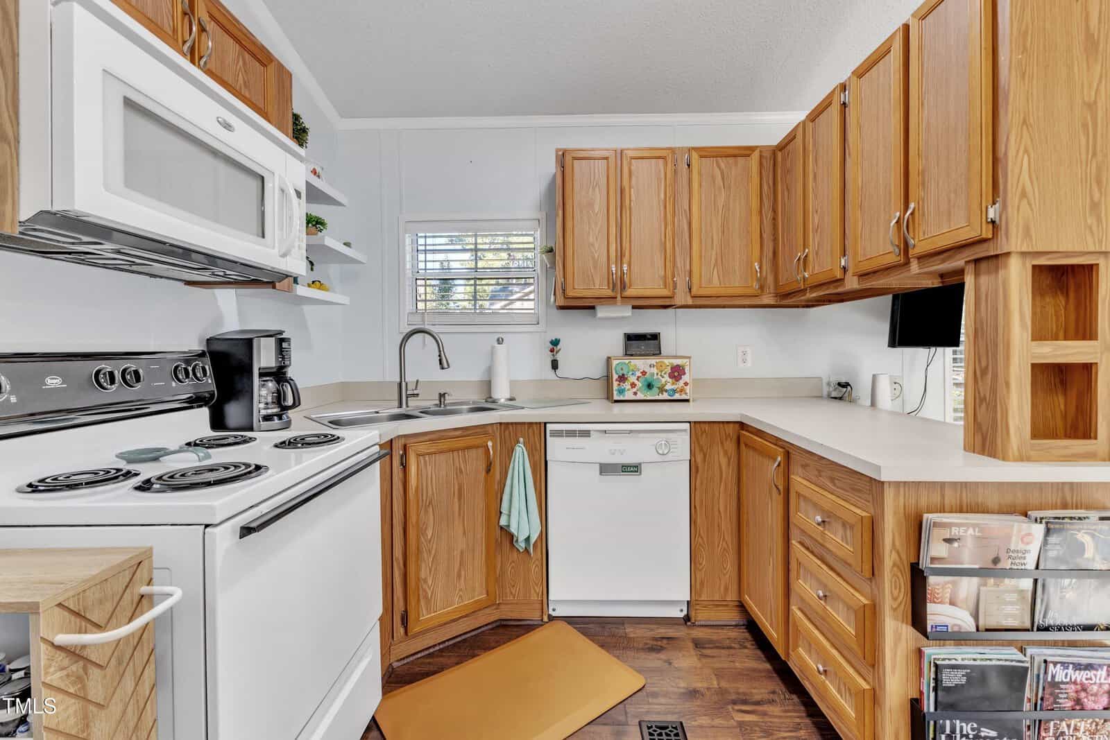Cozy kitchen with wood cabinets, white appliances including a stove, microwave, and dishwasher, a coffee maker, and a small window above the sink. Shelves hold books and magazines; sunlight streams through the window.