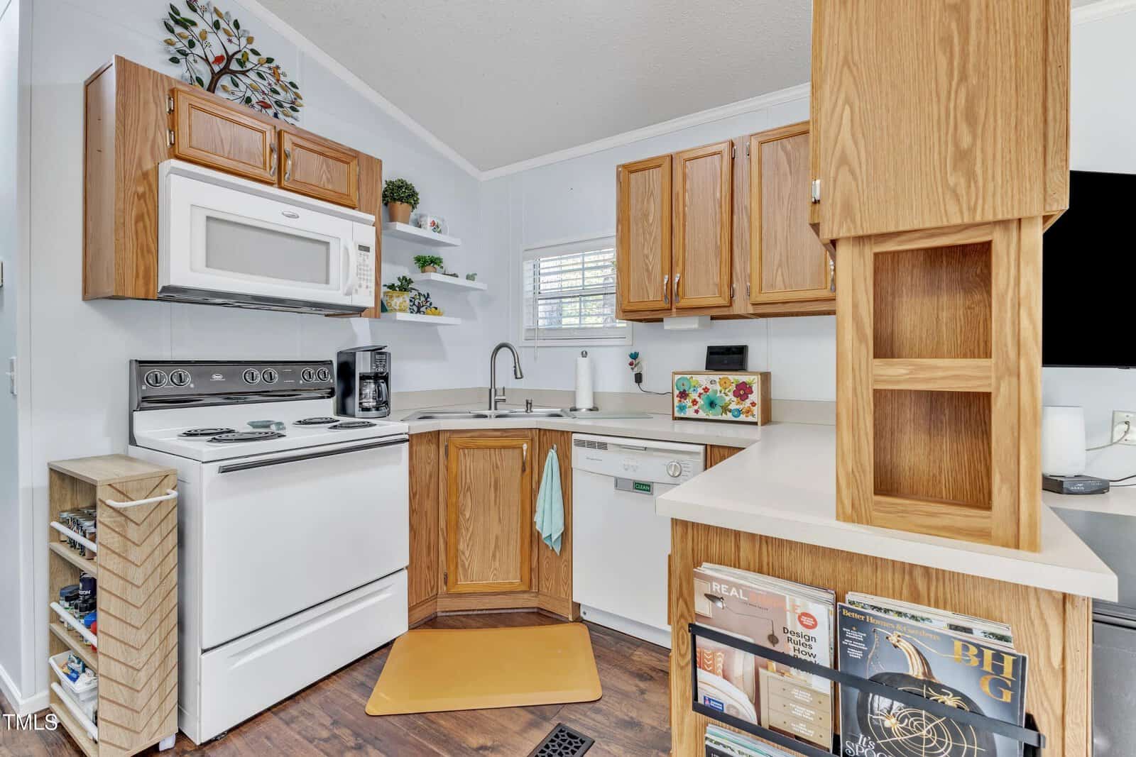 Small kitchen with wood cabinets, white appliances, a stove, microwave, dishwasher, and sink under a window. Shelves hold decor and a coffee maker. Records and magazines are stored on open shelves by the counter.