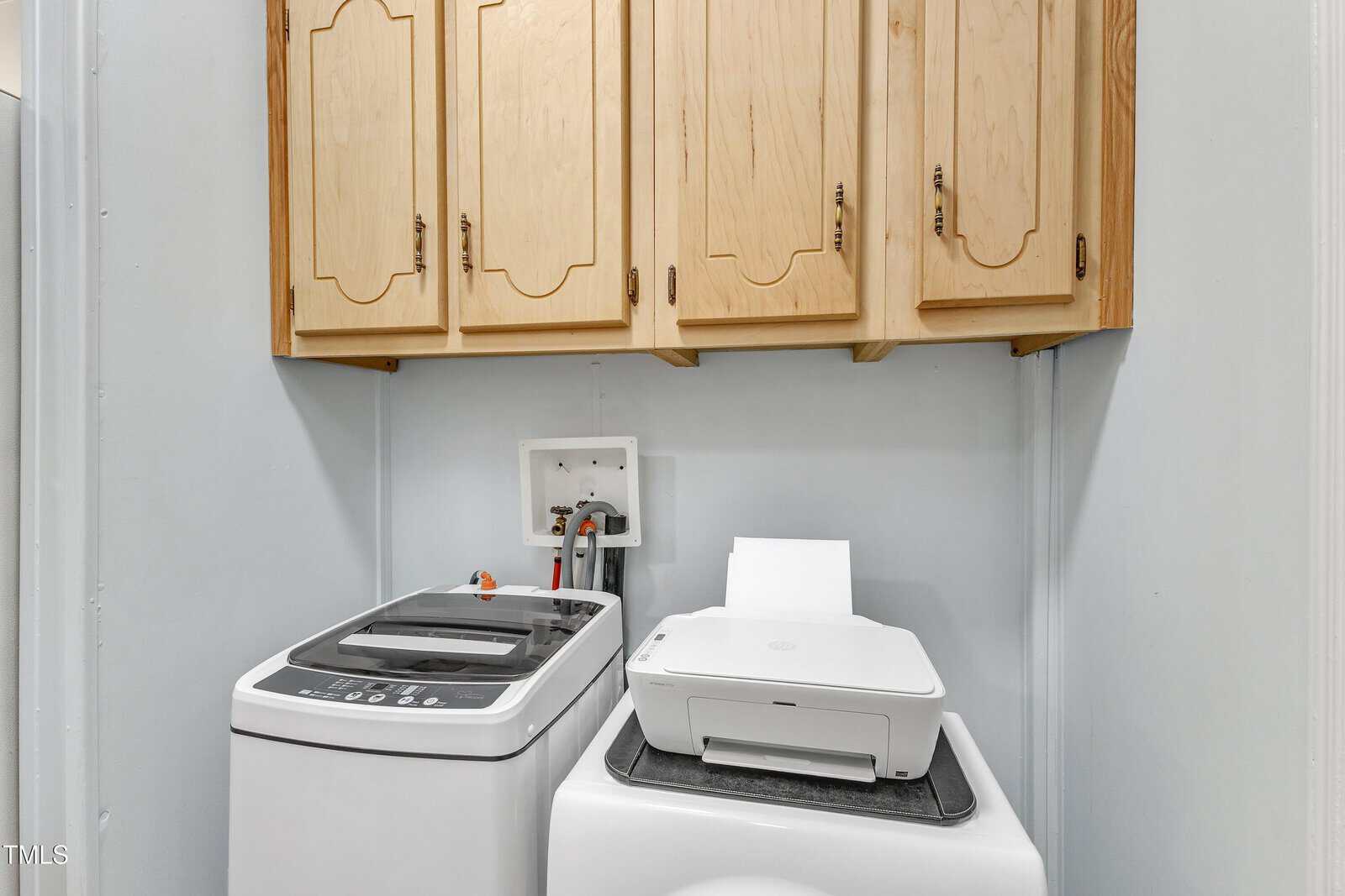 A laundry room with a top-loading washing machine, a front-loading dryer with a printer on top, wooden cabinets above, and white walls.