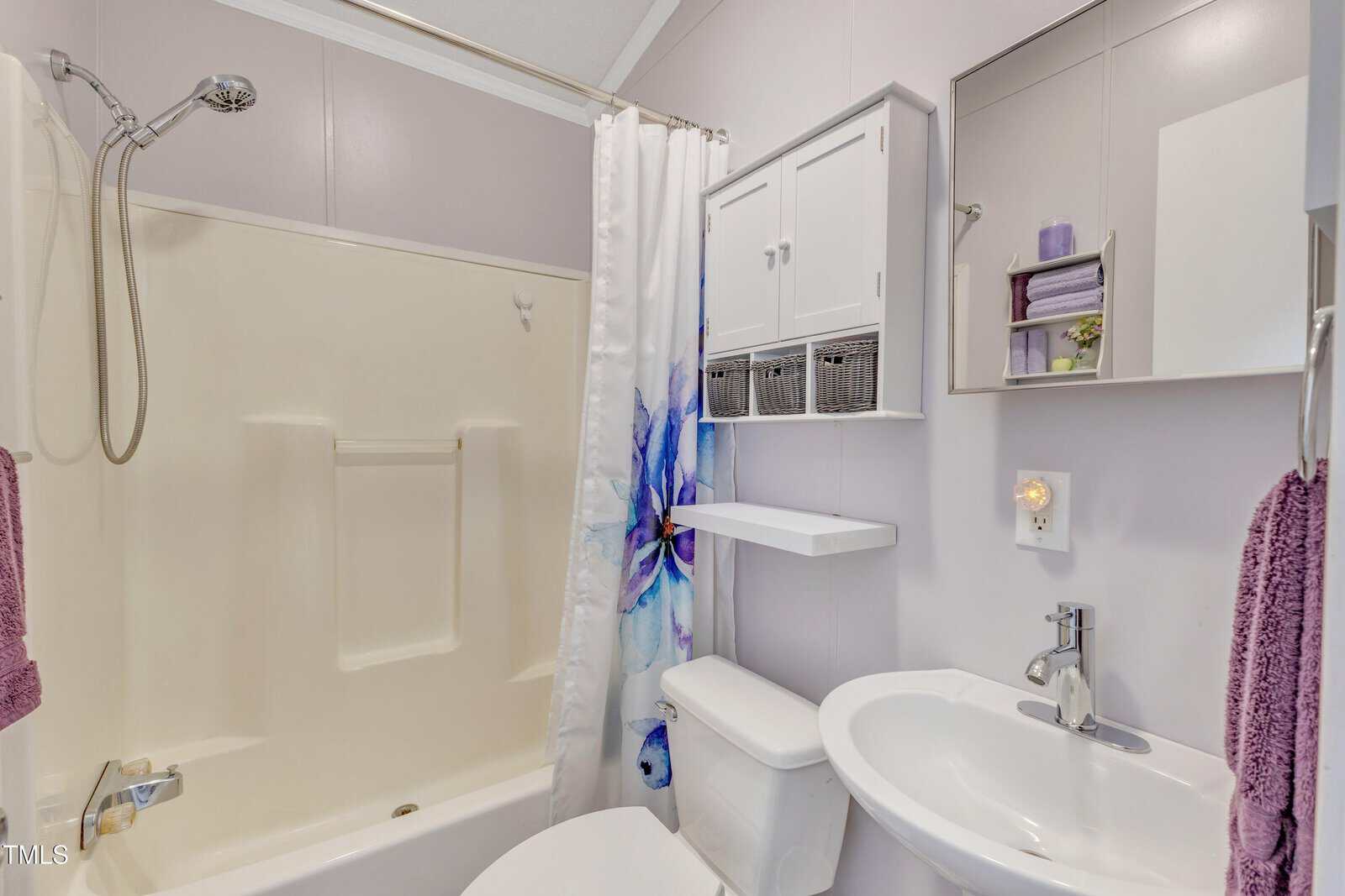 Small bathroom with a cream bathtub and shower, floral shower curtain, white toilet, pedestal sink, wall cabinet, shelves with towels, and a mirror above the sink. Purple and white color scheme.