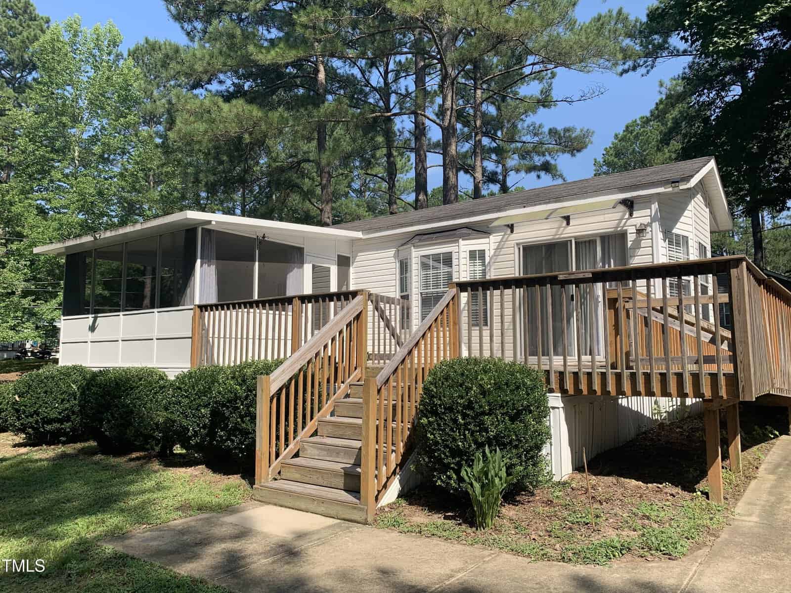 A white manufactured home with a screened porch, large sliding glass doors, and a wooden ramp and stairs. The home is surrounded by green shrubs, grass, trees, and is set in a wooded area on a sunny day.