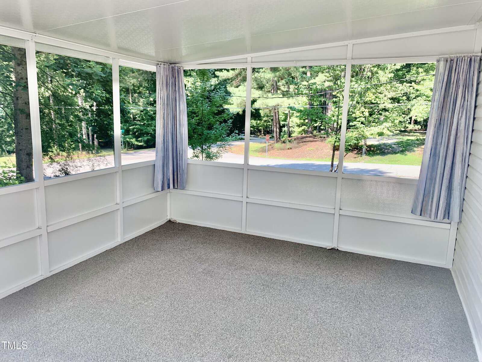 A bright, enclosed porch with screened windows, light blue curtains, and gray carpet flooring. Trees and greenery are visible outside, creating a peaceful, airy atmosphere.