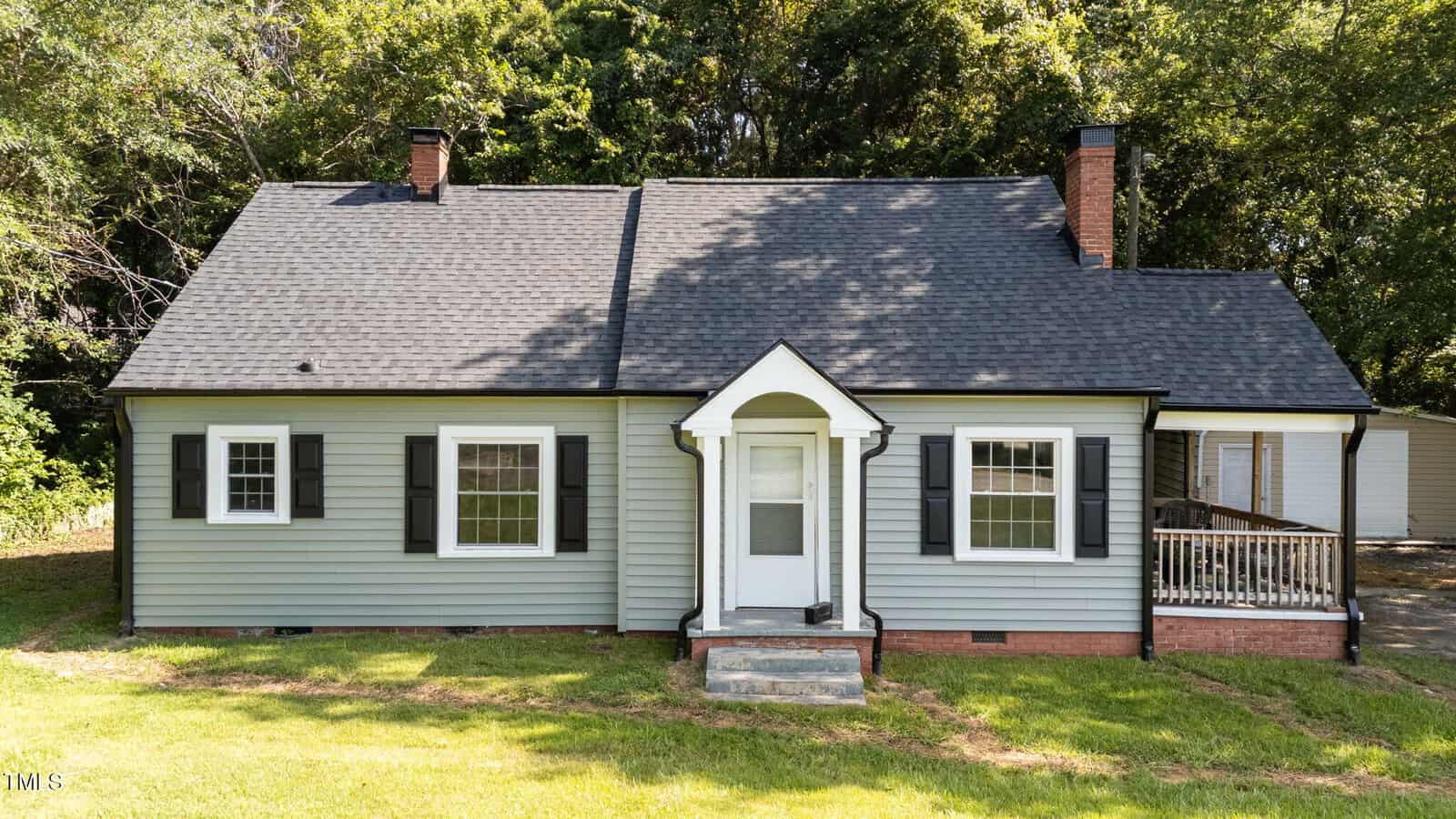A single-story house with gray siding, black shutters, two chimneys, and a small covered front porch sits at 205 Williamson Street in Louisburg. The house rests on a grassy lawn with trees in the background and features a ramp leading to a side entrance.