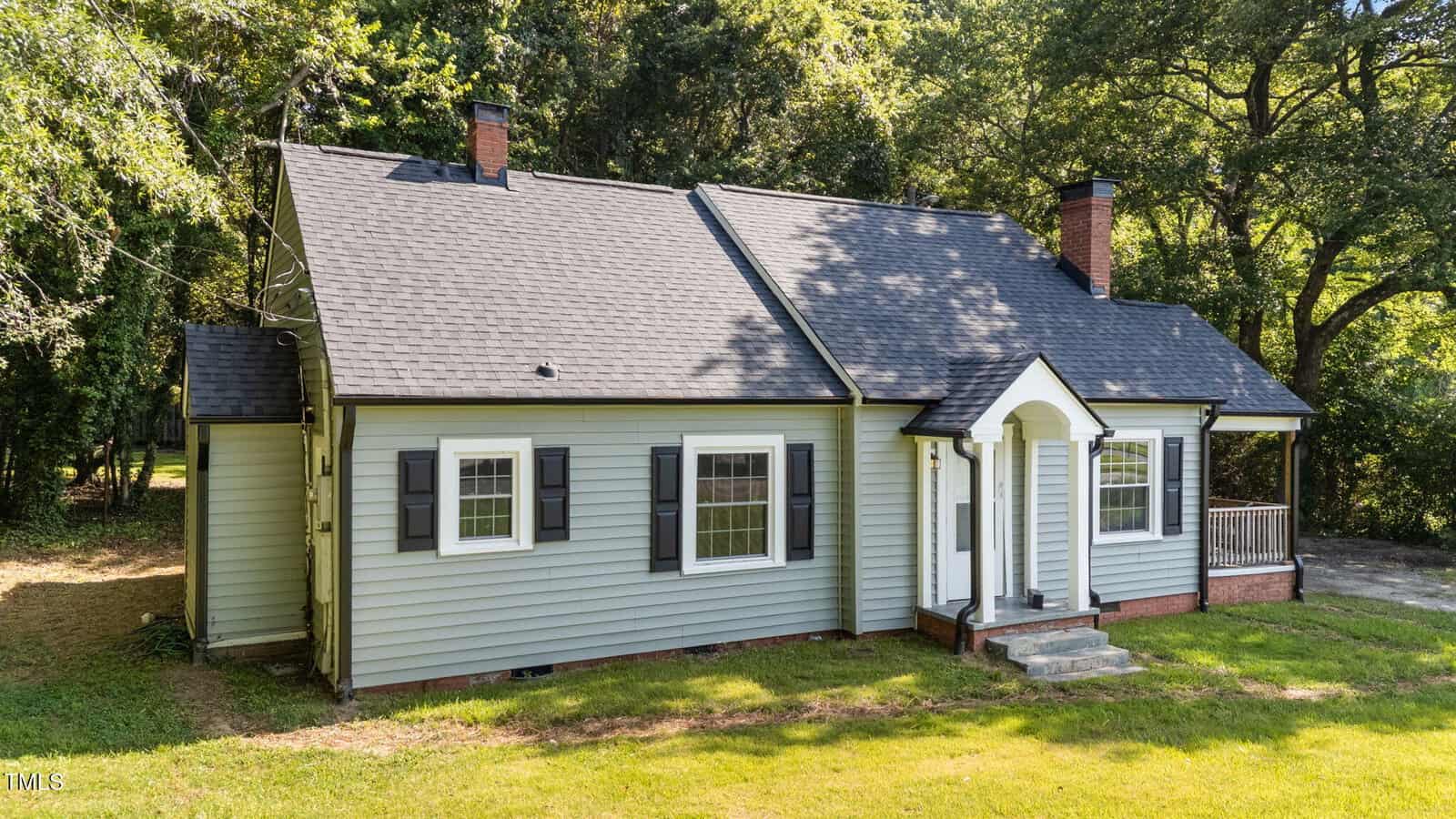 Light gray single-story house with black shutters, a dark roof, two chimneys, a small front porch with white columns, and steps leading to the front door. Surrounded by grass, trees, and shade.