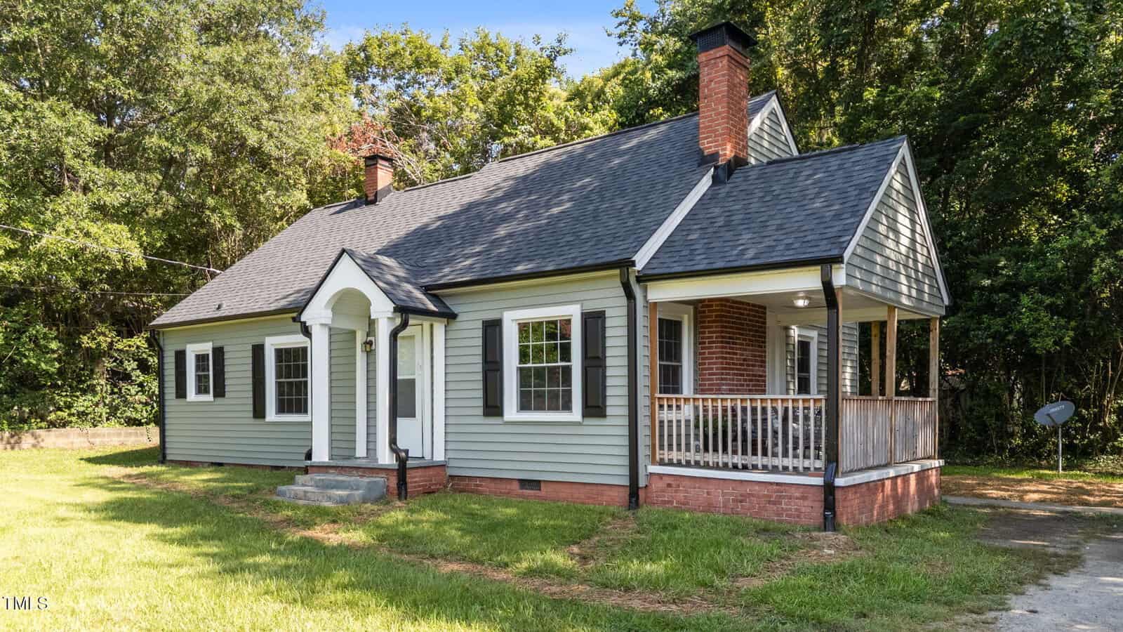 A single-story, light green house with black shutters, a dark shingled roof, two brick chimneys, and a covered porch with wooden railings. The front entrance has an arched portico. The house is situated on a lawn with trees in the background.
