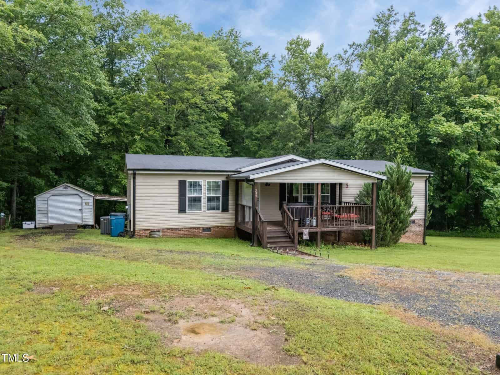 A single-story manufactured home with beige siding, black roof, and front porch sits on a grassy lot with a gravel driveway; trees and a small shed are visible in the background.