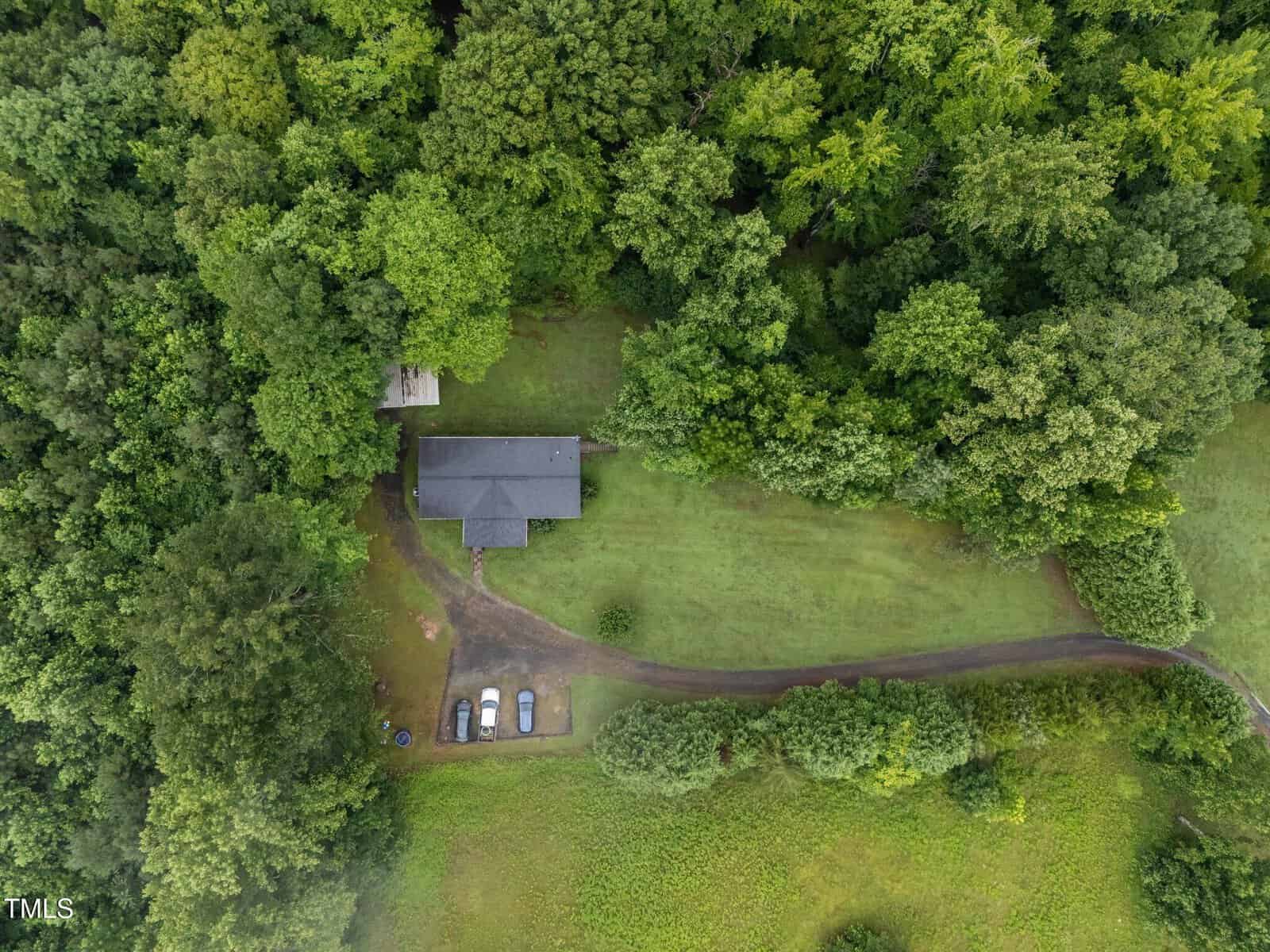 Aerial view of a house surrounded by dense trees, with a driveway leading to the house and four parked cars on a grassy area. The property is mostly bordered by forest.