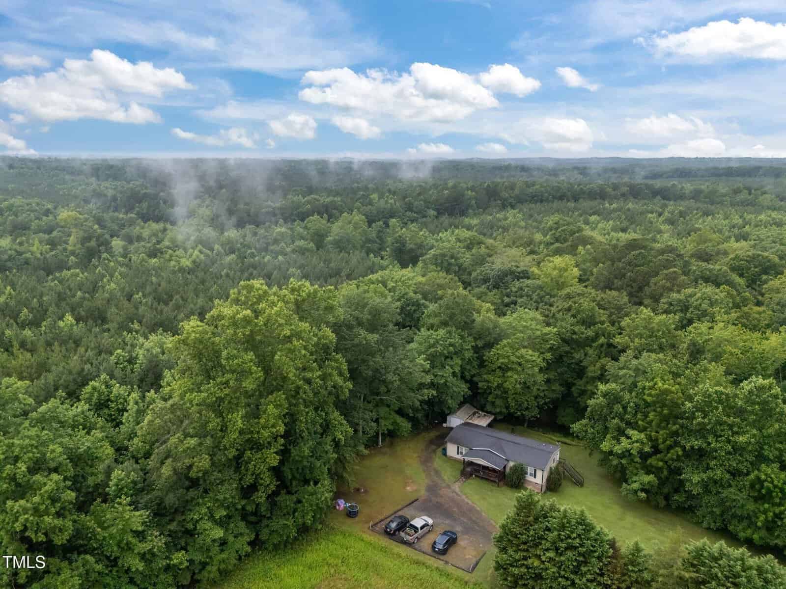 Aerial view of a house at 544 Duke Drive, Rougemont, surrounded by dense green forest. A gravel driveway leads to the house, with four parked cars nearby. The sky is partly cloudy, and mist drifts among the distant trees.