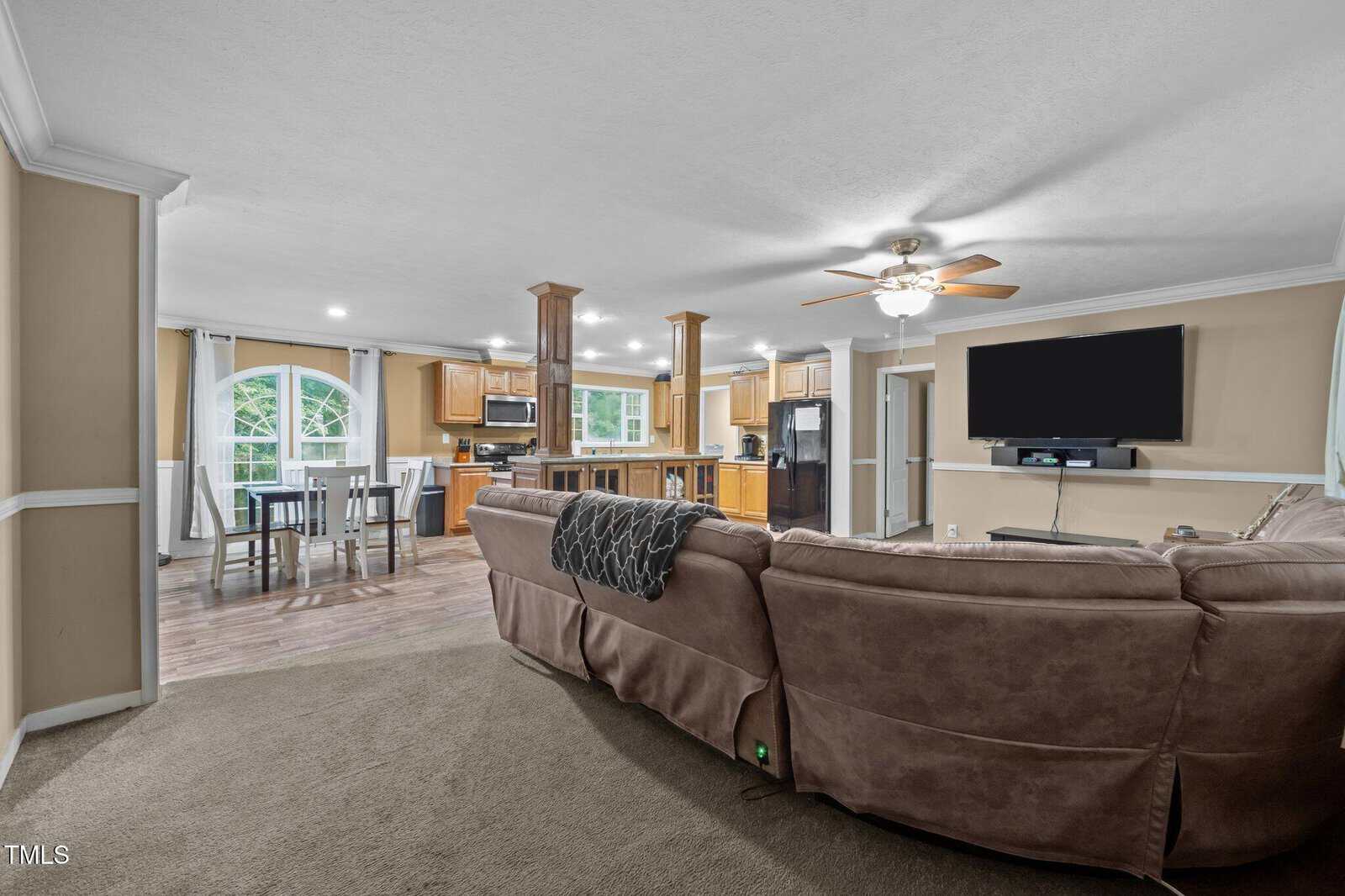 A spacious open-concept living area with a large sectional sofa, wall-mounted TV, and ceiling fan. The kitchen with wood cabinets and a dining area with large windows are visible in the background.