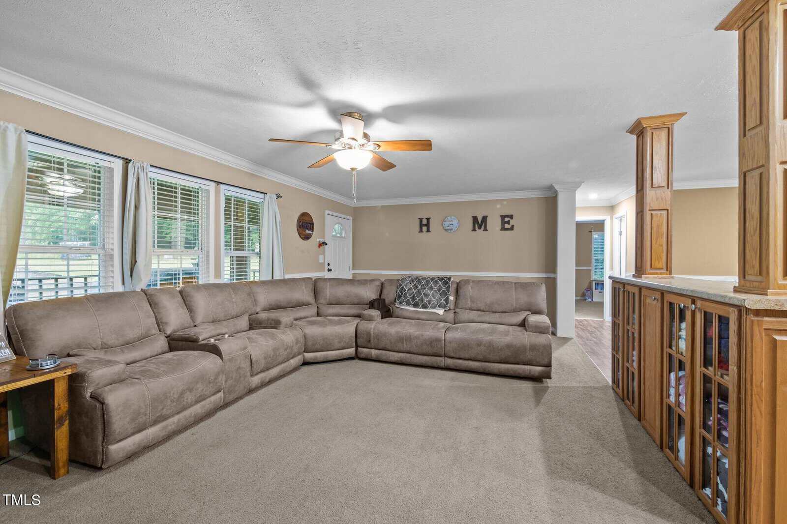 A living room with a large beige sectional sofa, a wooden side table, carpeted floor, ceiling fan, and windows with white curtains. The word "HOME" is displayed on the far wall above a doorway.